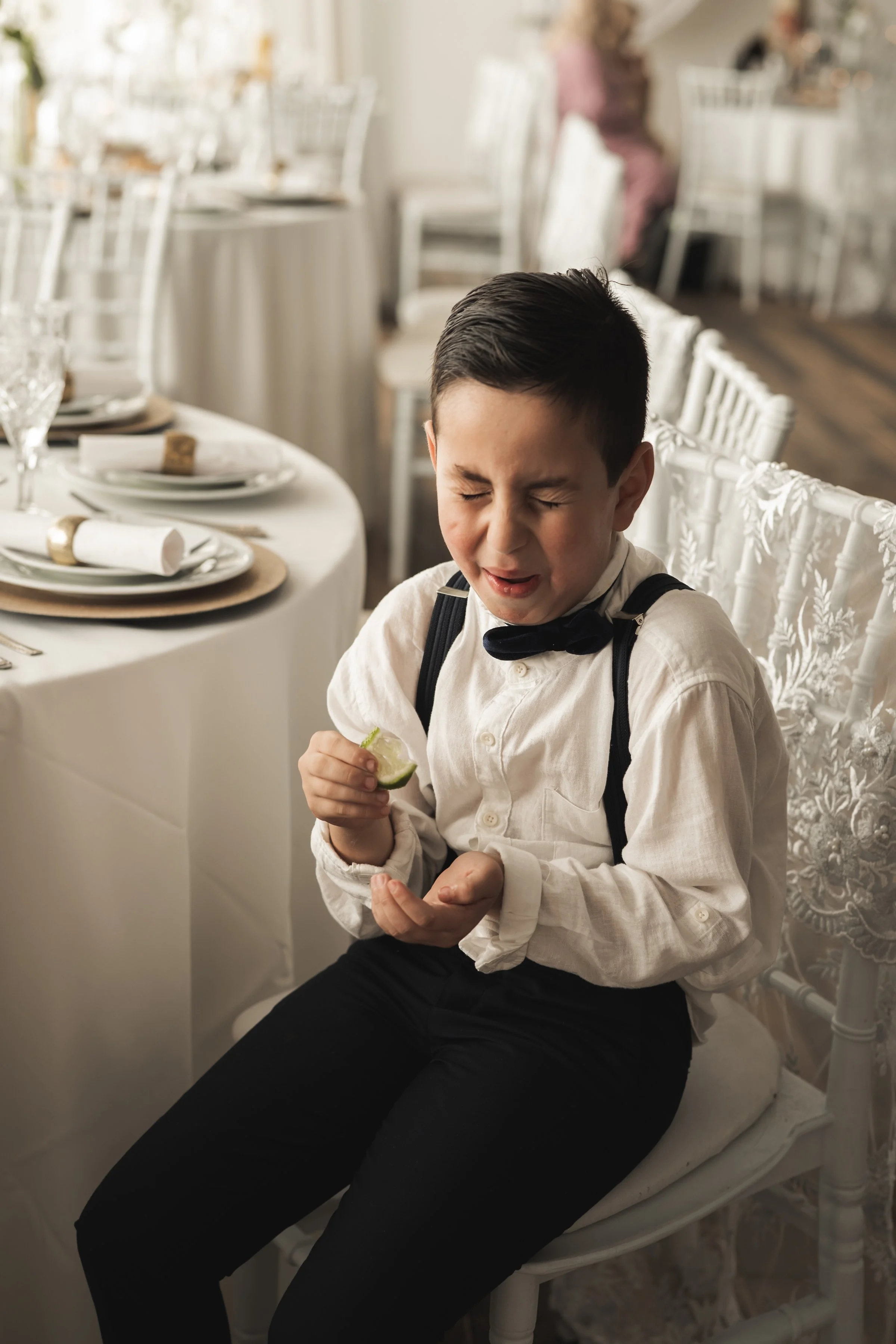 Een jongen in een wit overhemd met een strikje zit aan een gedekte tafel in een feestzaal, met een pijnlijke uitdrukking en een schouder gevuld met een rugzak.