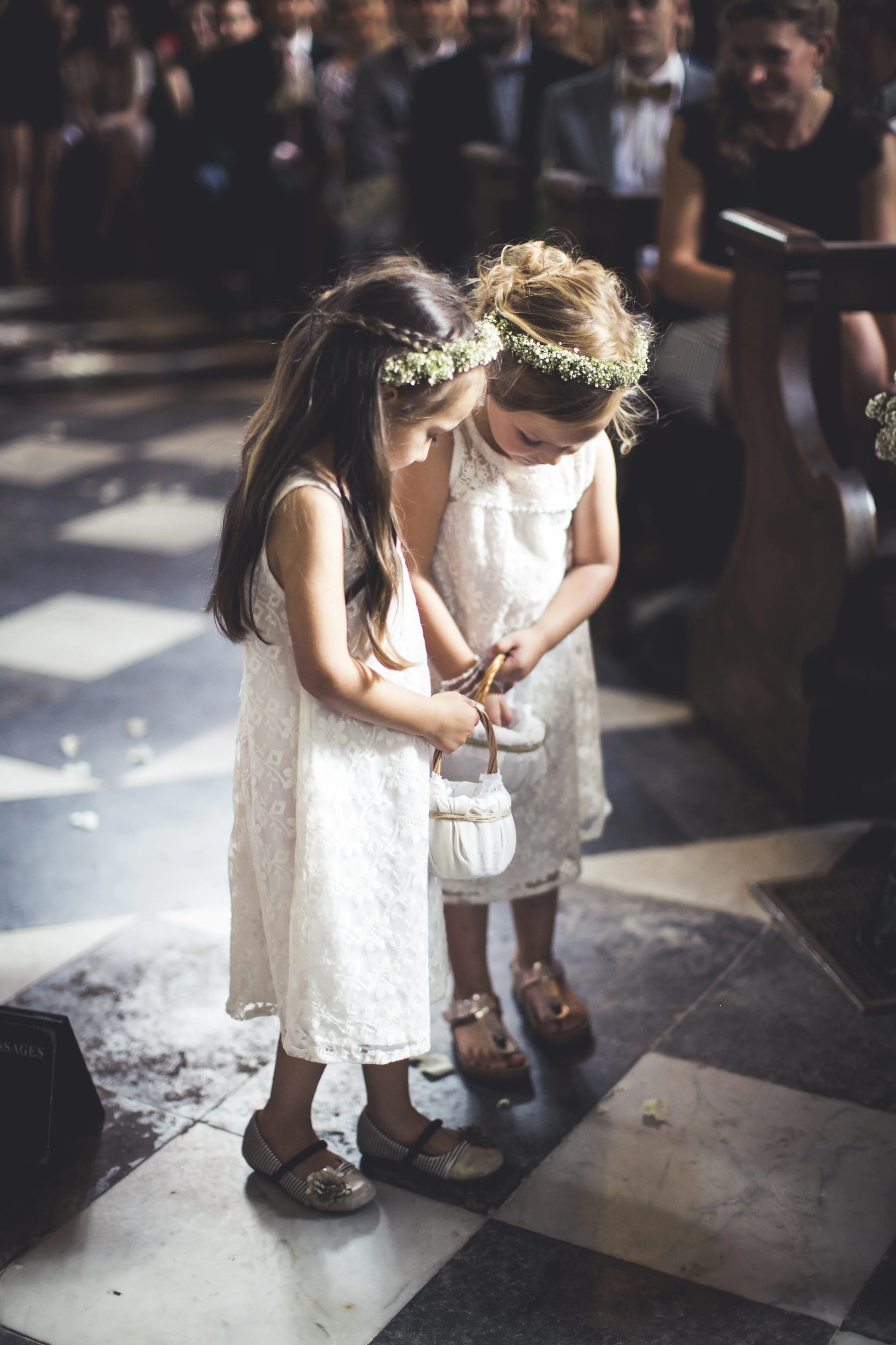 Twee jonge meisjes in witte jurkjes met bloemenkransen op hun hoofd in een kerk tijdens een ceremonie, met hun hoofden gebogen en handen bij elkaar.