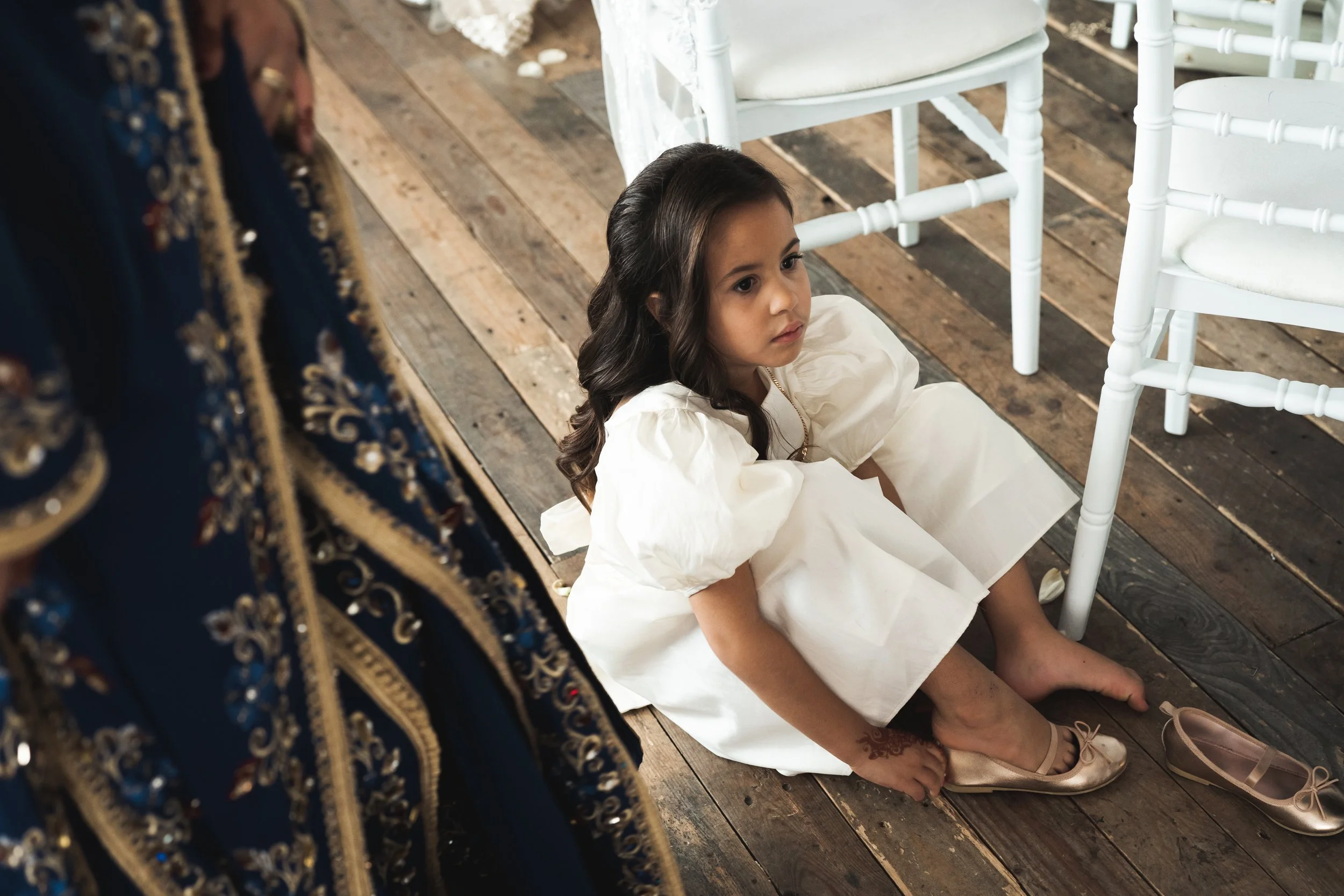 Een jonge meid in een witte jurk zit op de houten vloer, haar schoenen te snel uitdoen, met schone schoenen naast haar.
