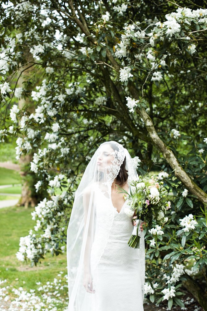 Een bruid in een witte trouwjurk met sluier terwijl ze een bouquet vasthoudt, stond onder een bloeiende boom met witte bloemen in een tuin.