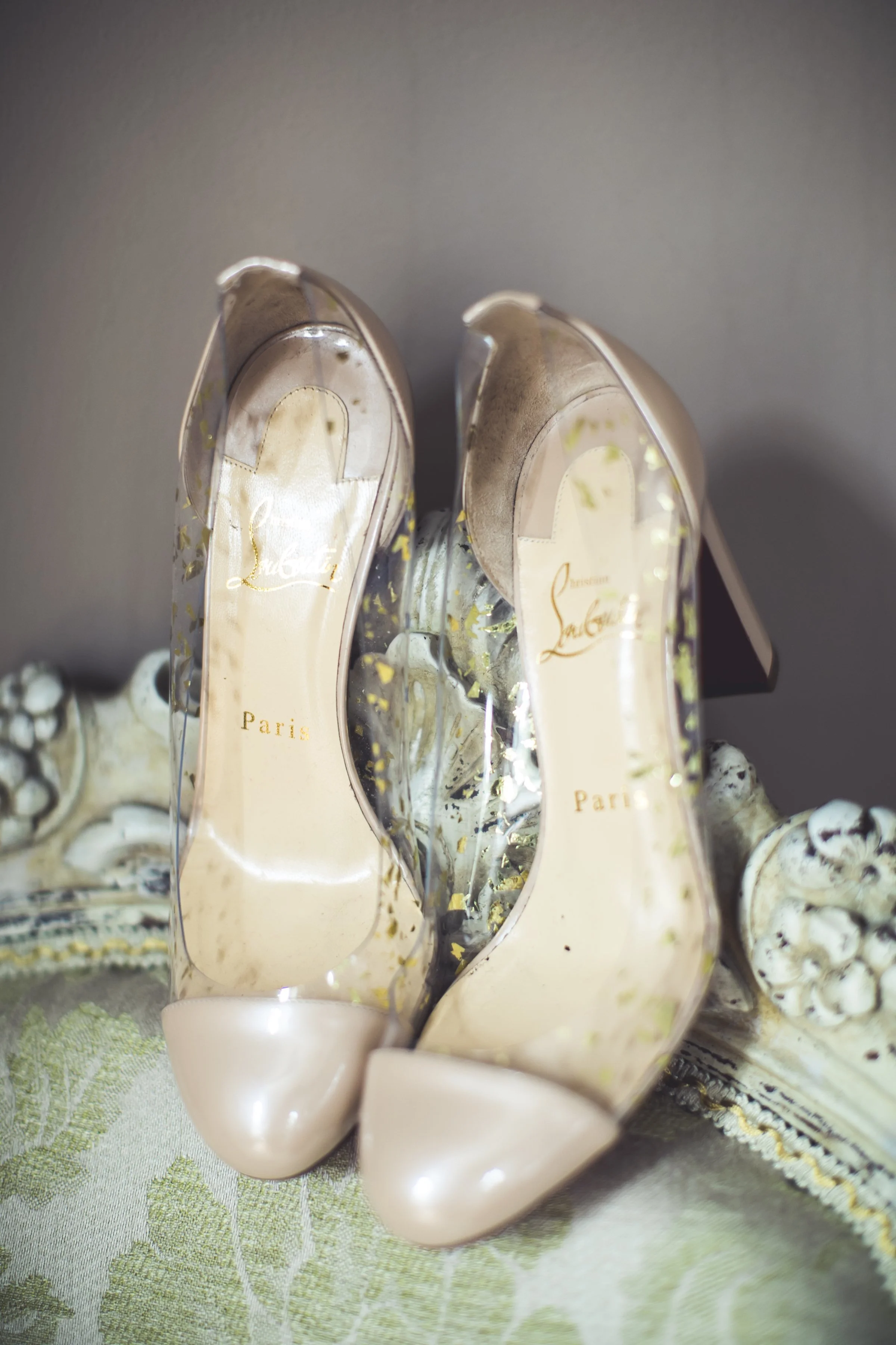 Transparante hoge hakken met gouden details en tekst 'Paris' aan de binnenzool, geplaatst op een sierlijke ornamentele rand op een tafel.