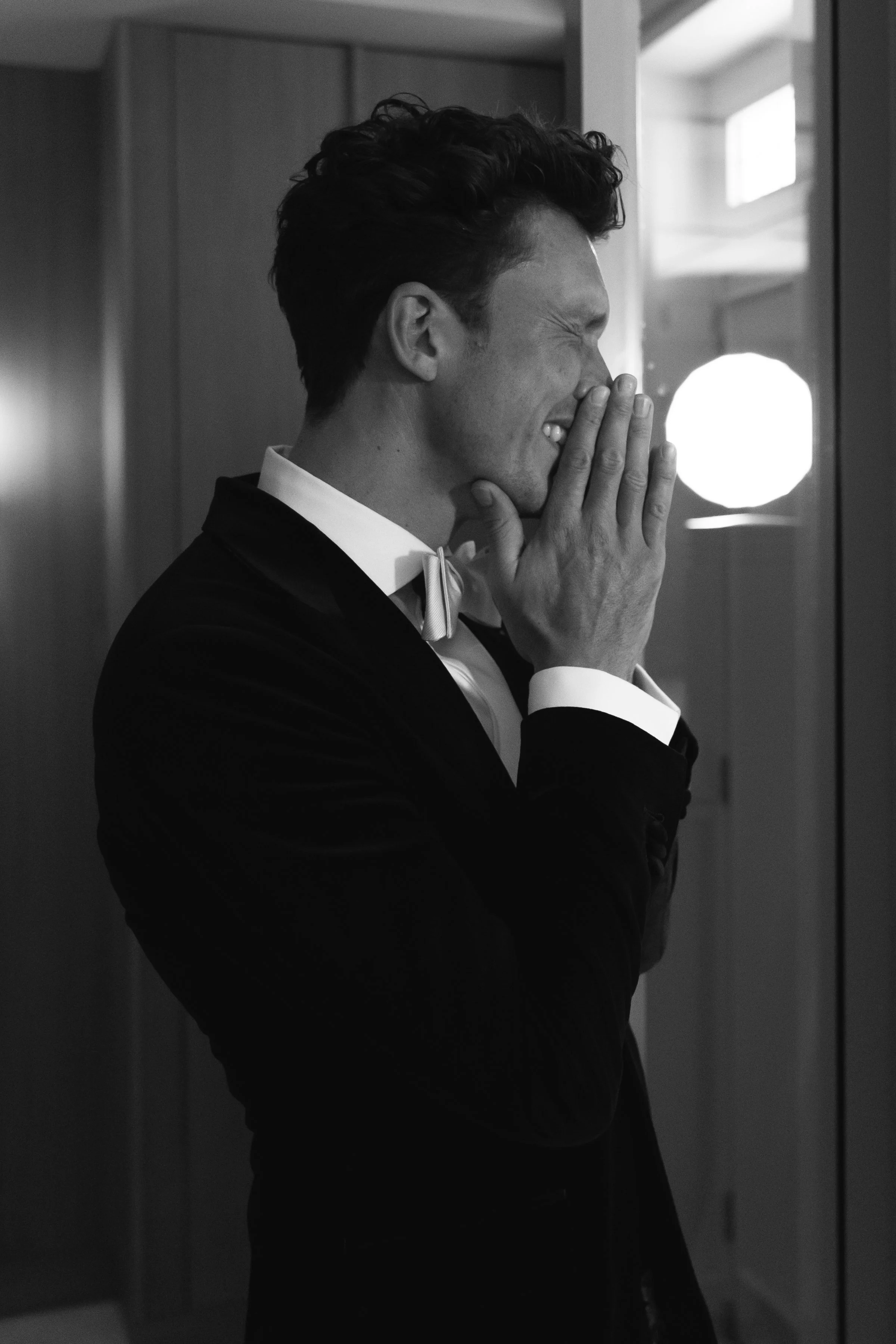 Man in smoking, lachend met handen voor mond, binnen, zwart-wit.