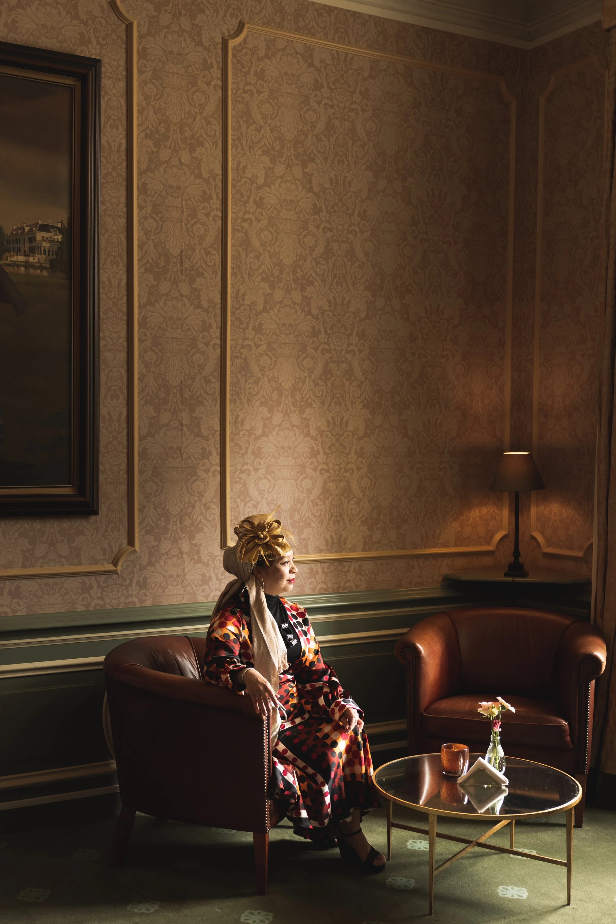 Vrouw in een kleurrijk patroonjurk en een opvallende hoed zit in een elegante kamer met vintage inrichting, naast een tafel met bloemen.
