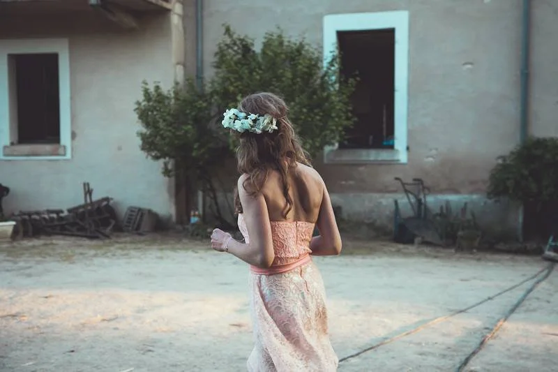 Een vrouw met een bloemenkrans op haar hoofd, gekleed in een lichtroze jurk, staat buiten voor een verouderd gebouw.