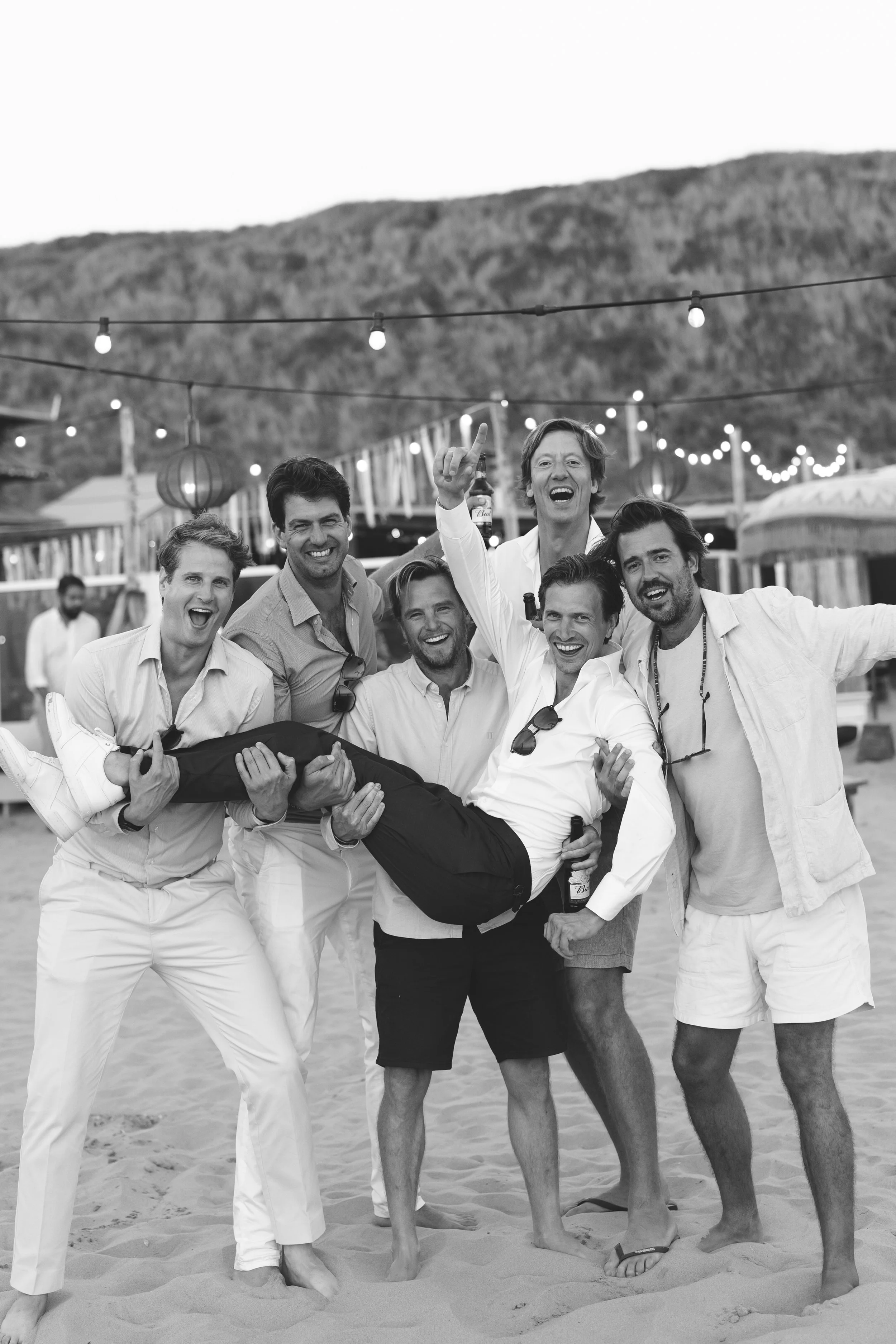 Vrouw wordt lachend door zes mannen op het strand gedragen tijdens feest, met lichtslingers en lampionnen op de achtergrond.