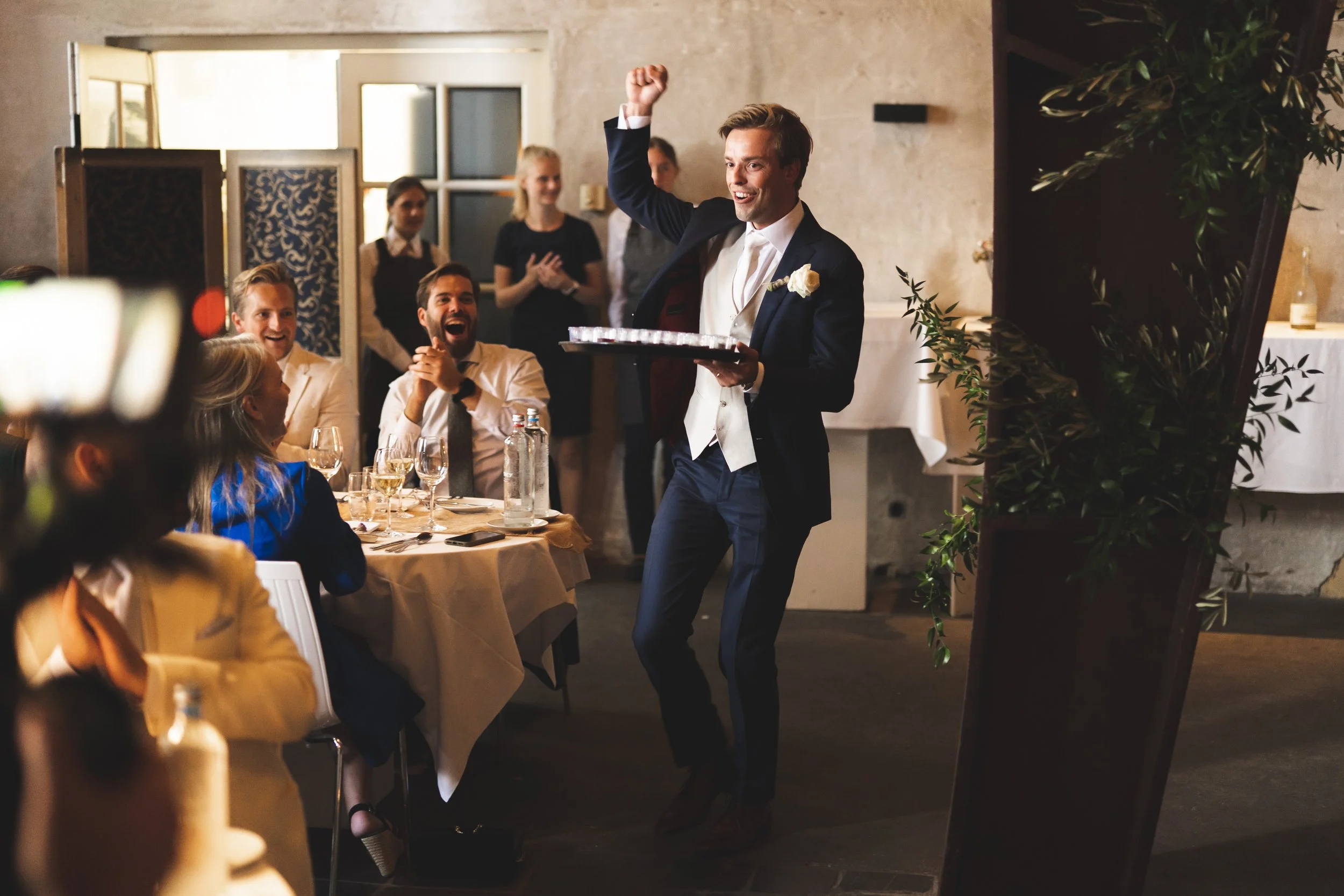 Man in een smoking die een toast uitbrengt op een bruiloft, omringd door lachende gasten.