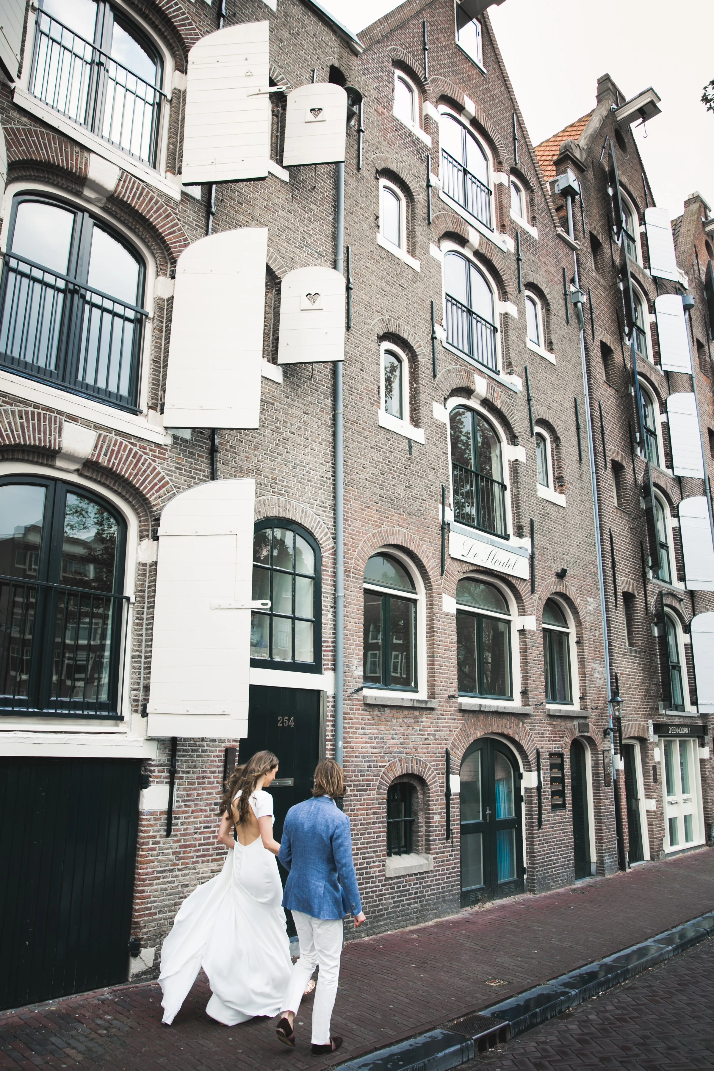 Een man en een vrouw in een witte trouwjurk lopen samen langs een gebouw met bakstenen muren en open witte luiken.