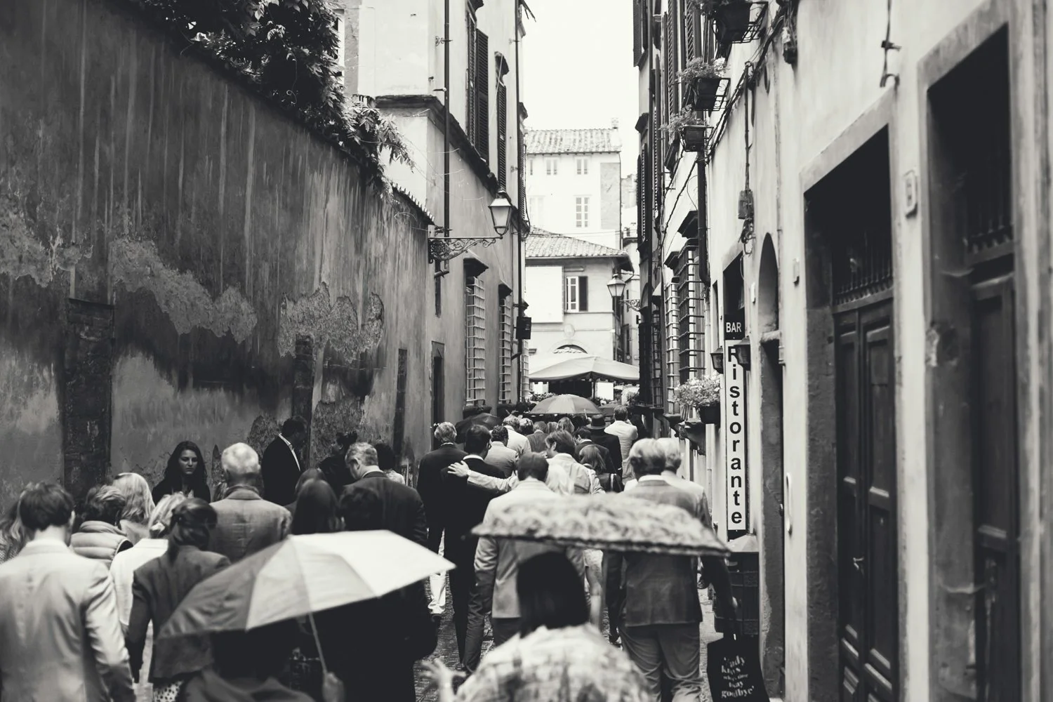 Een zwart-wit foto van een drukke straat met veel mensen die met paraplu's lopen, omringd door oude gebouwen met hoekige ramen en gevels in een stad.