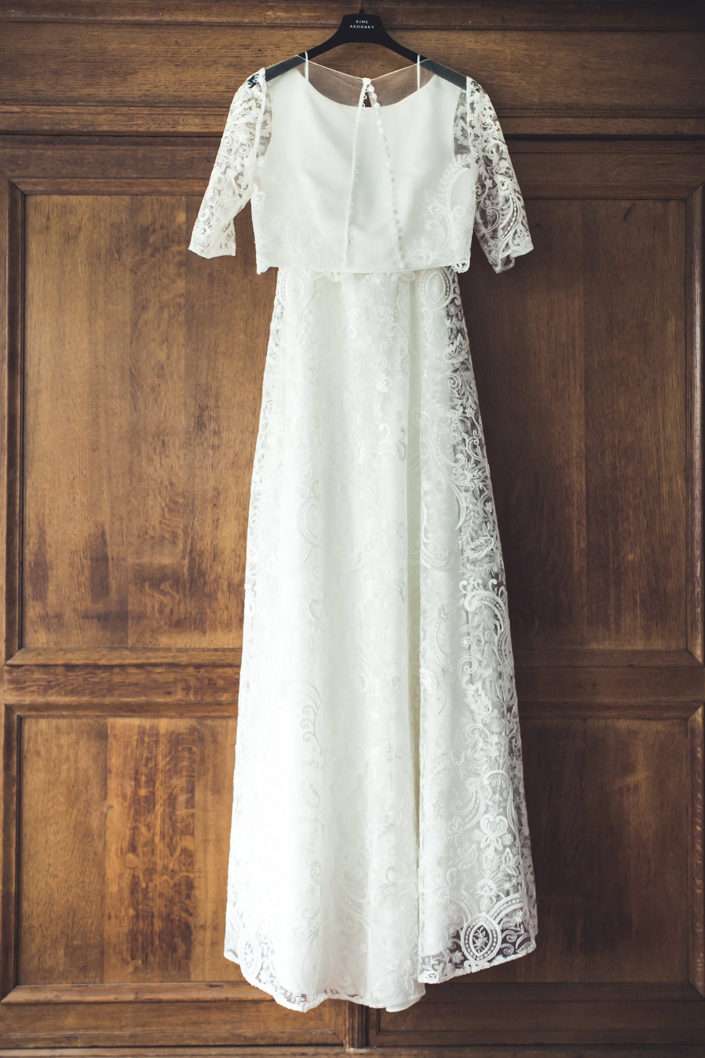 Witte trouwjurk met kanten top en jasje, hangend aan een kapstok op een houten muur.