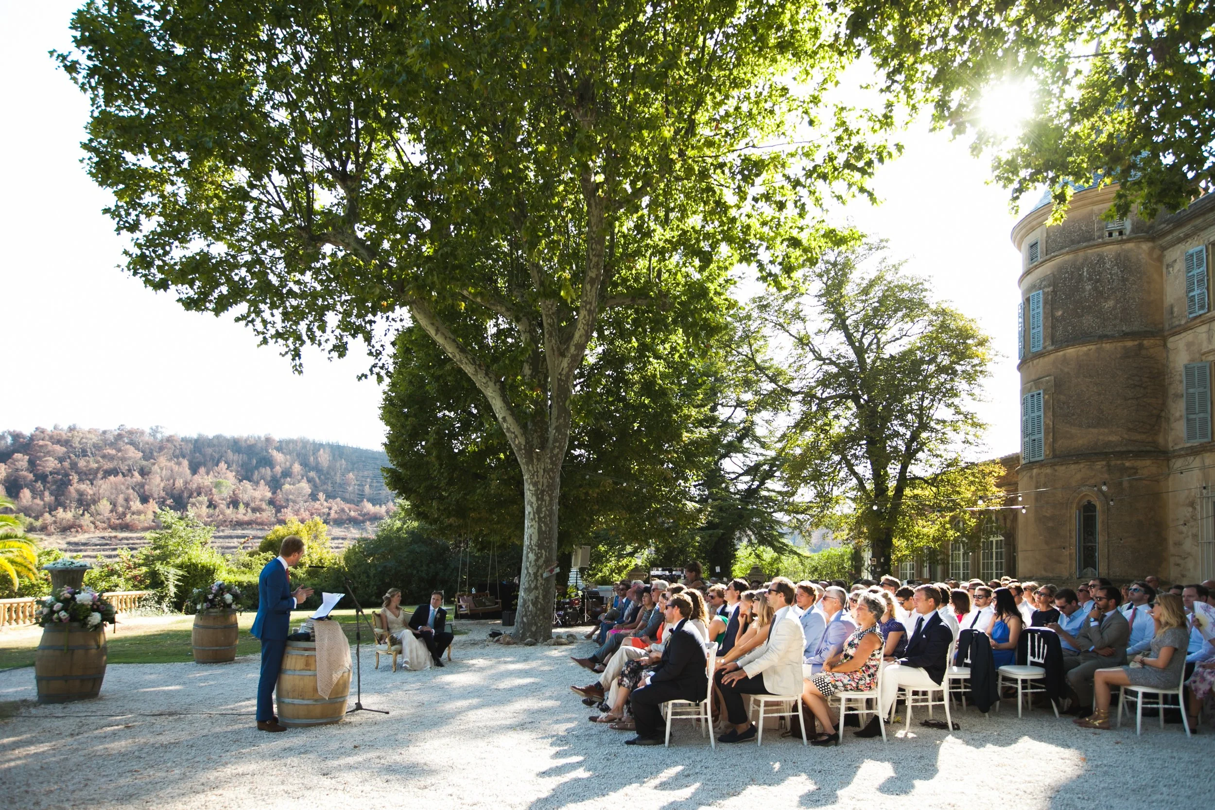 Mensen die getuige zijn van een bruiloftsceremonie buiten onder een grote groene boom met zonlicht in de achtergrond.