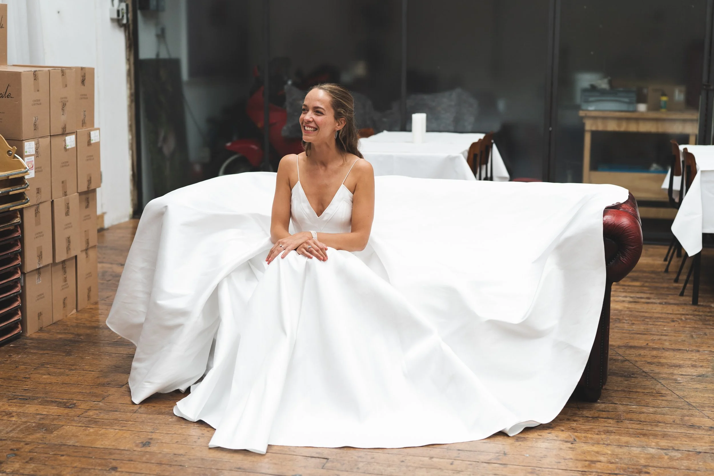 Een vrouw in een witte jurk zit glimlachend op een bank in een ruimte met houten vloer en papieren dozen aan de linkerkant.