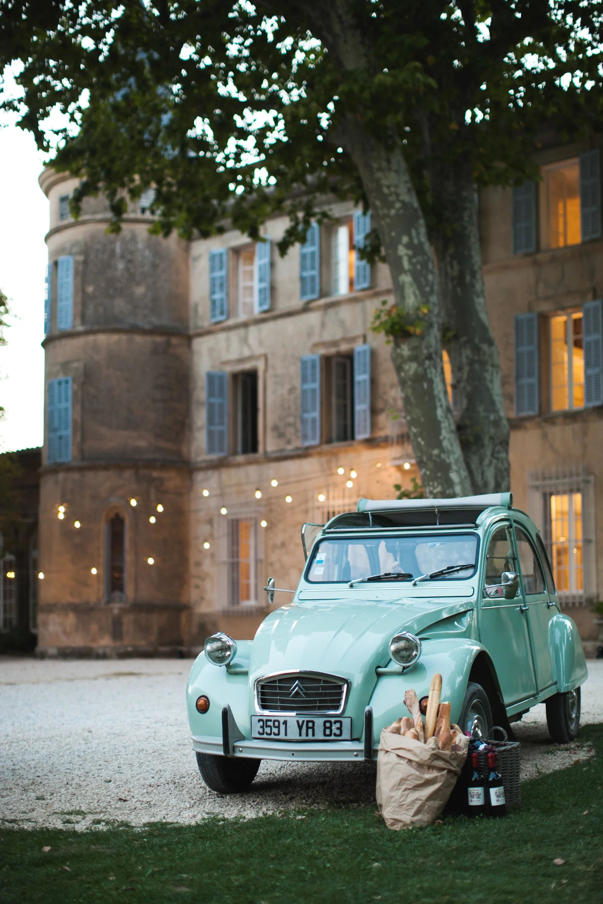 Een lichtblauwe Citroën 2CV auto geparkeerd voor een oud kasteel, met een tas met baguettes en twee flessen wijn op de grond in de buurt.