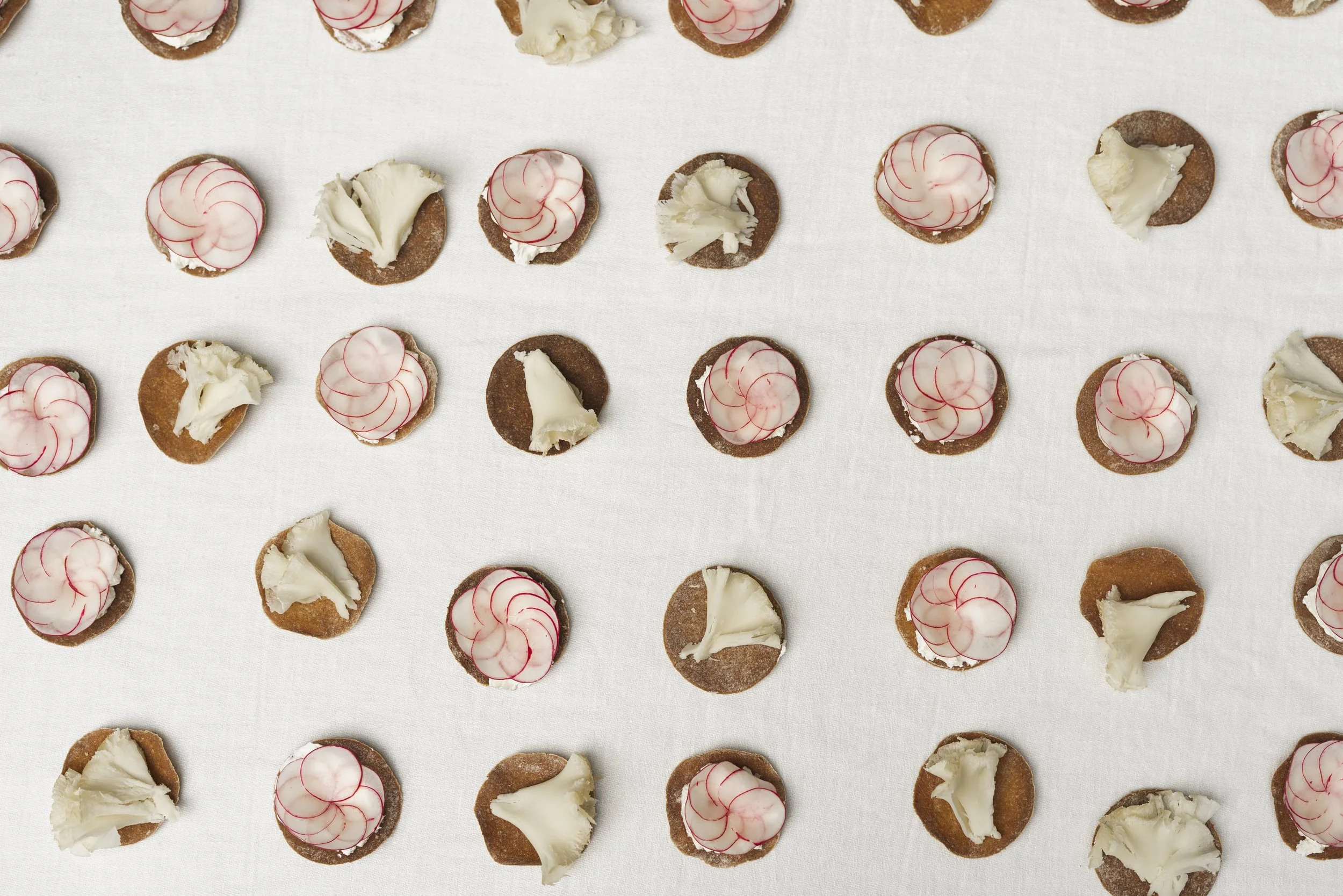 Rijen met kleine rondjes met witte en rode decoratie, en stukjes witte chocolade op een witte tafel. De decoraties lijken op kleine koekjes met topping.