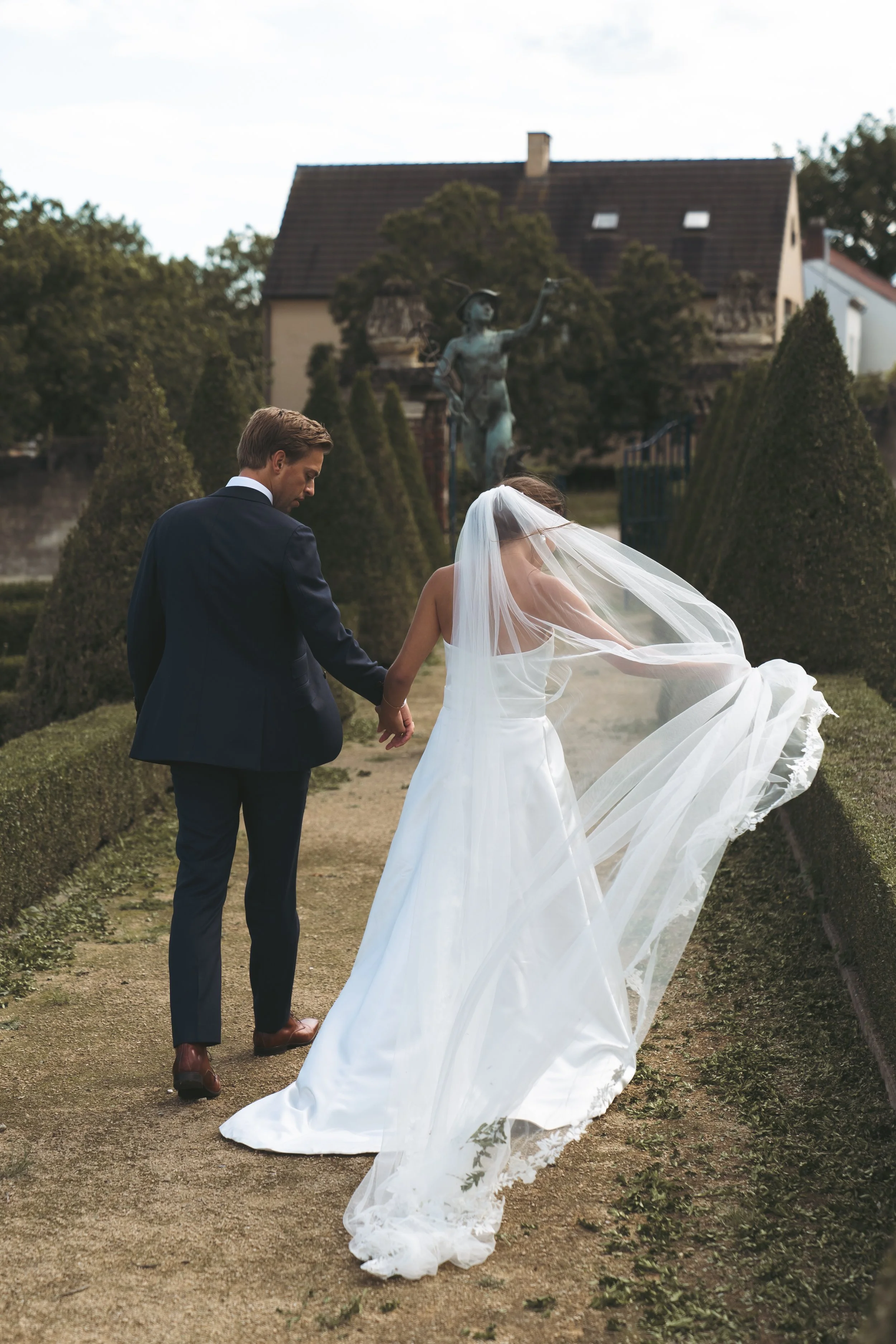 Een pasgetrouwd paar wandelt hand in hand door een tuin, de bruid draagt een lange witte jurk en een sluier, de bruidegom in een donker pak, met op de achtergrond een standbeeld en een huis.