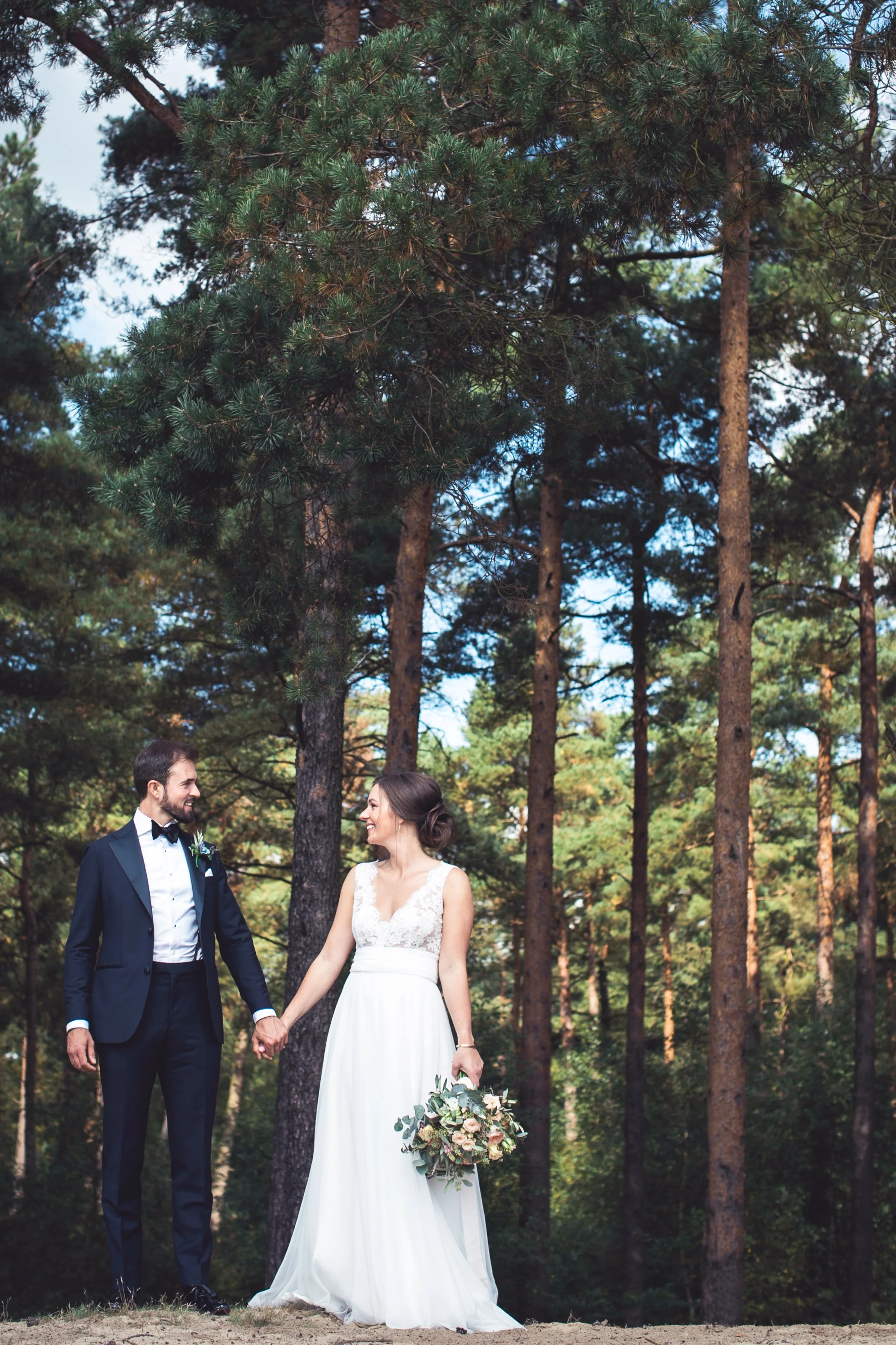 Een bruid en bruidegom staan hand in hand in een bos, lachend naar elkaar. De bruid draagt een witte trouwjurk en houdt een boeket bloemen. De bruidegom draagt een blauwe tuxedo en een strik.