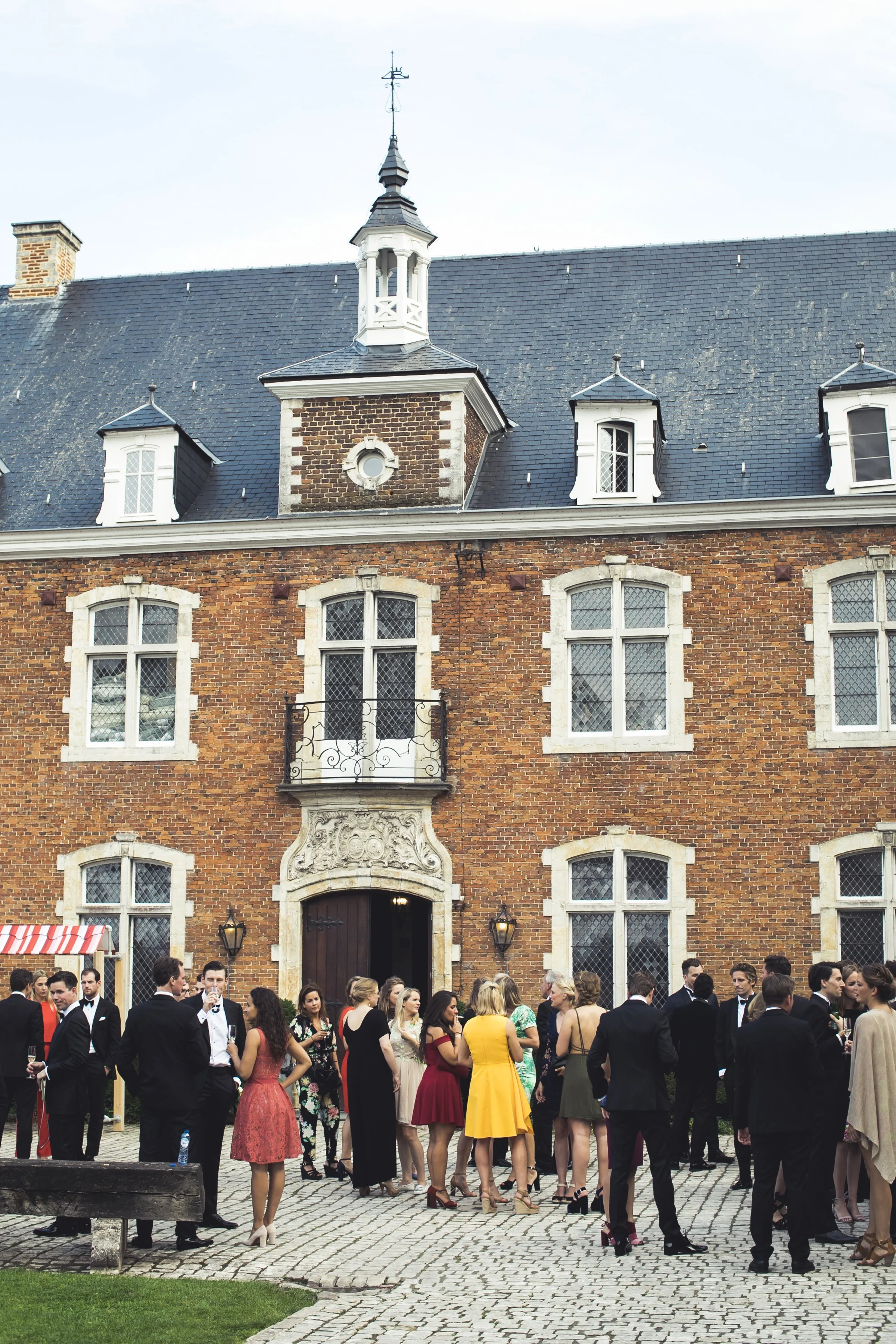 Mensen in formele kleding, waaronder mannen in tuxedo's en vrouwen in jurken, verzamelen zich buiten een historische stenen gebouw met grote ramen en een omgangswoning. Het lijkt op een feest of receptie.