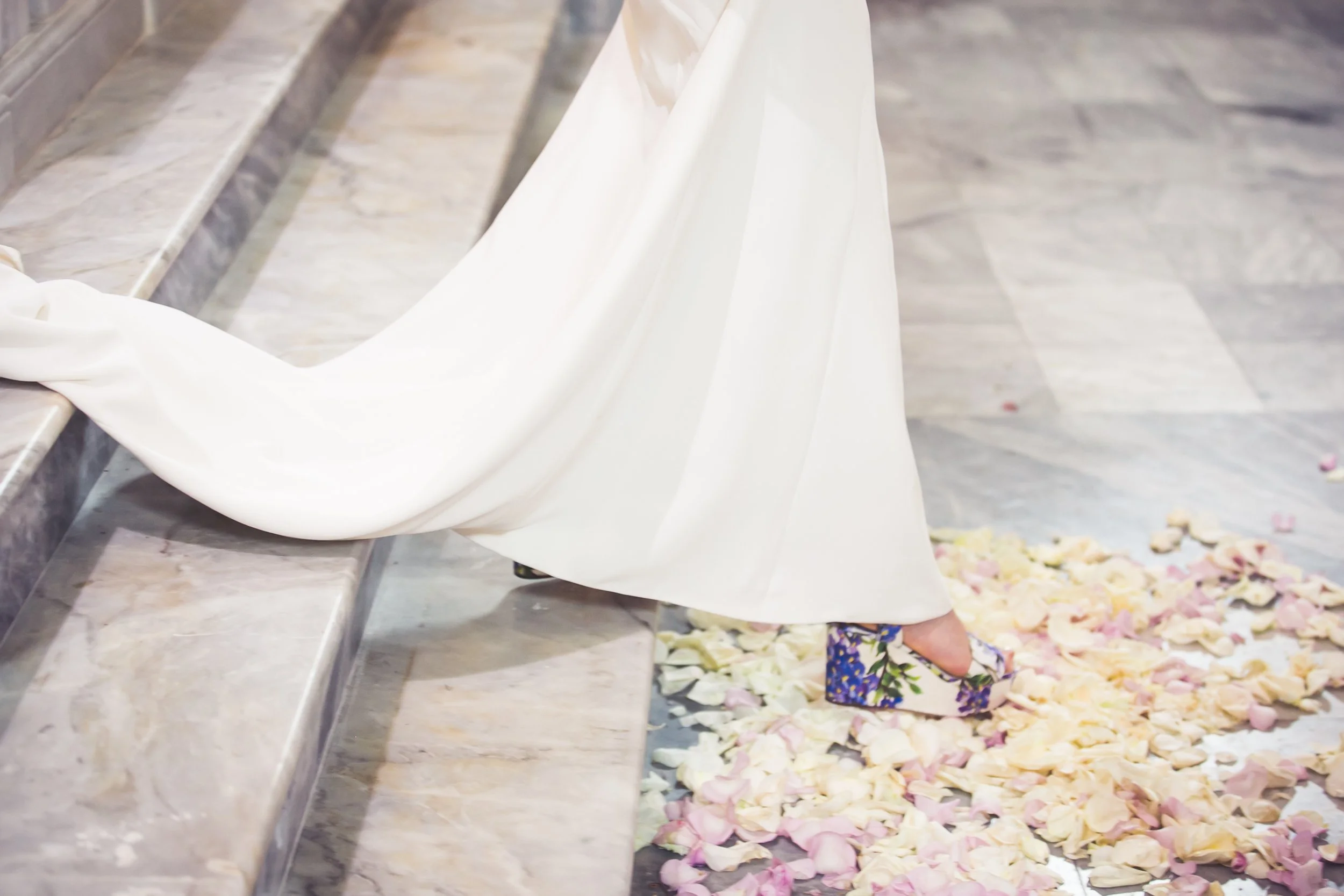 De zoomer in op de onderkant van een bruidssluier en een elegante schoen met een bloemenpatroon op een stenen vloer en rozenblaadjes op de grond.