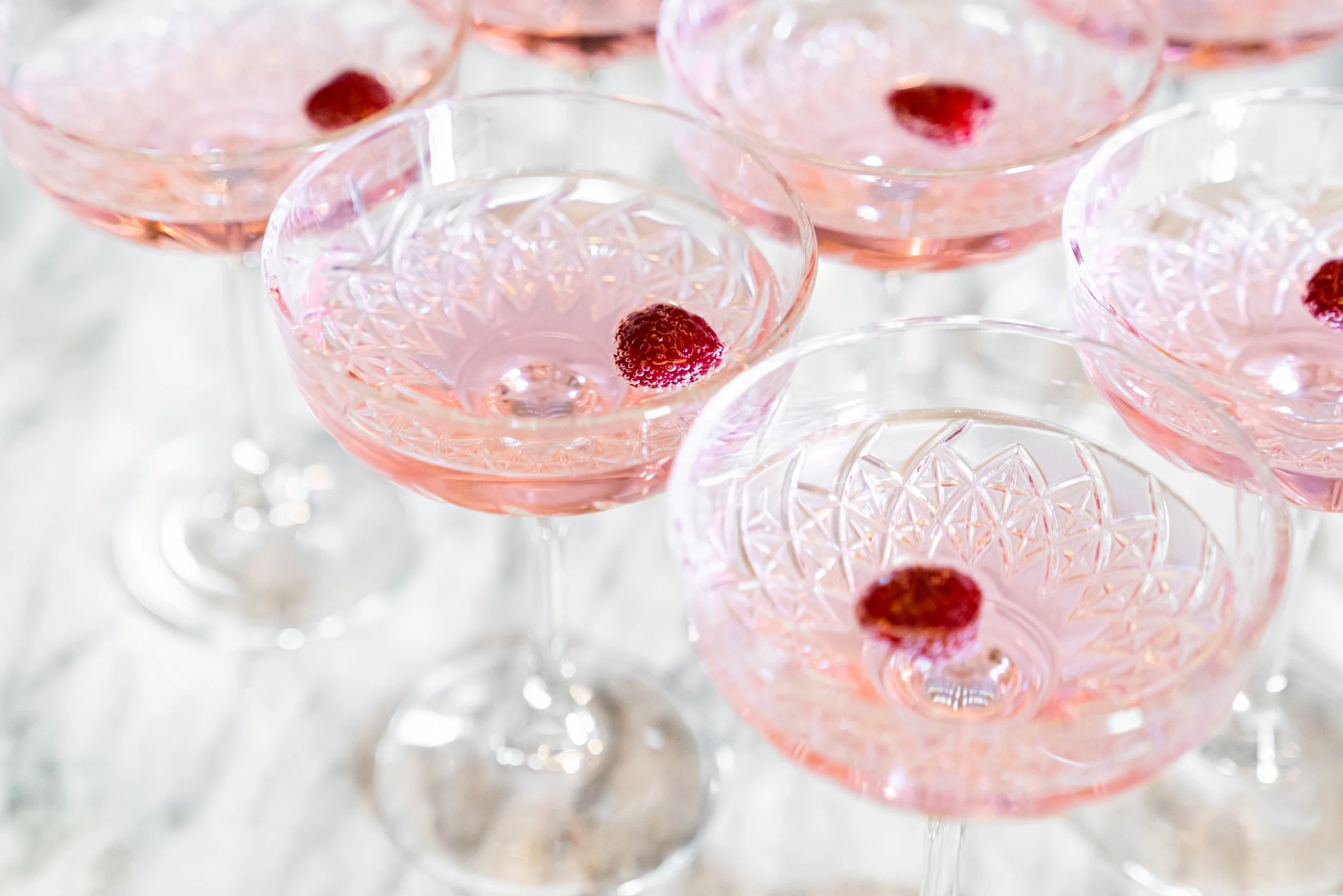 Verschillende champagne-coupes gevuld met een roze drank, elk met een framboos als garnering.