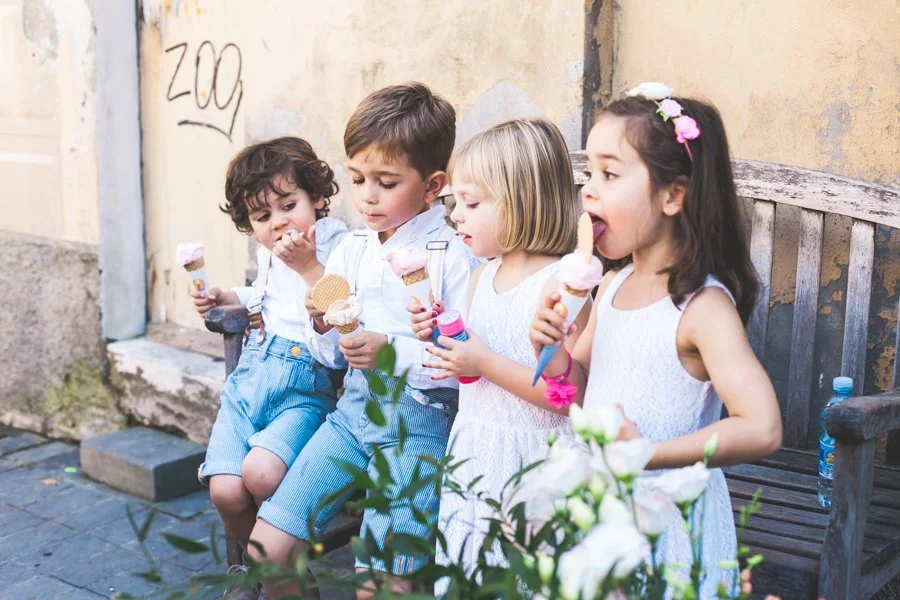 Vijf kinderen die ijsjes eten, zittend op een bankje tegen een verweerde muur.