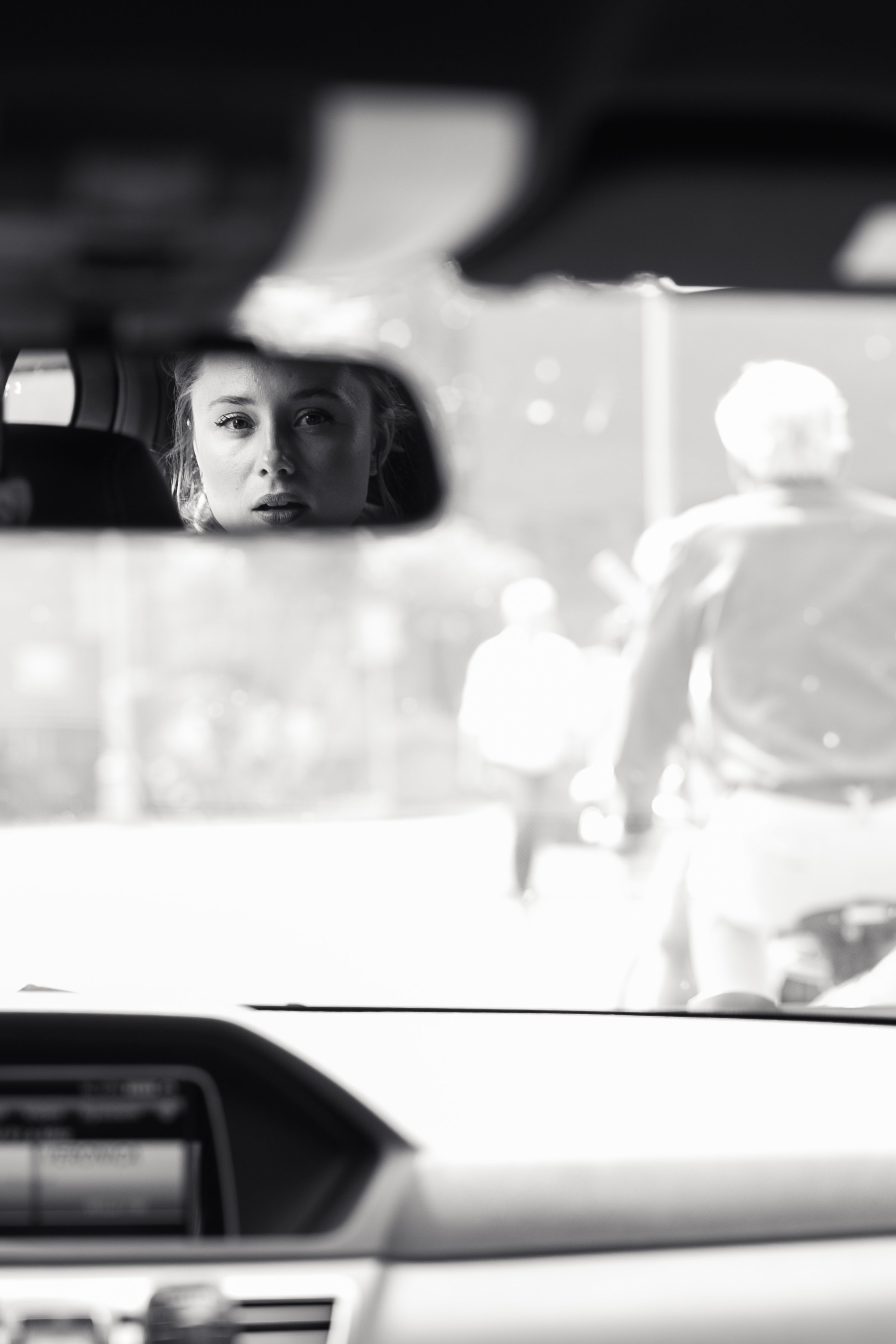 Een zwart-wit foto van een vrouw die in de achteruitkijkspiegel van een auto kijkt, met een persoon op de fiets op de achtergrond.