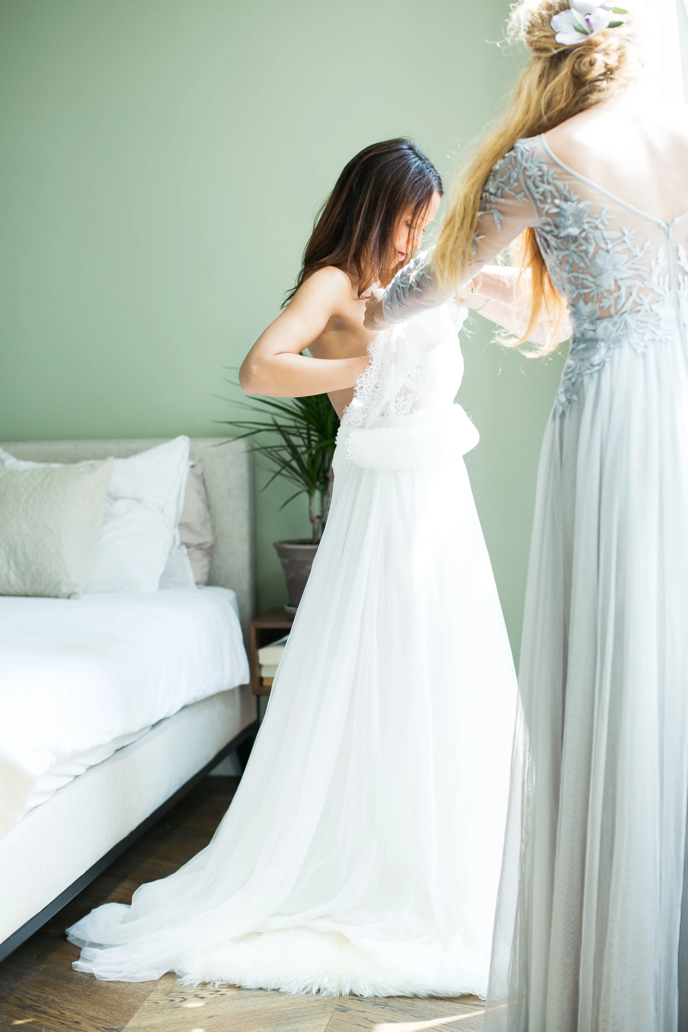 Vrouw krijgt een witte trouwjurk aangetrokken in een lichte kamer met groene muur en een bank met kussens.