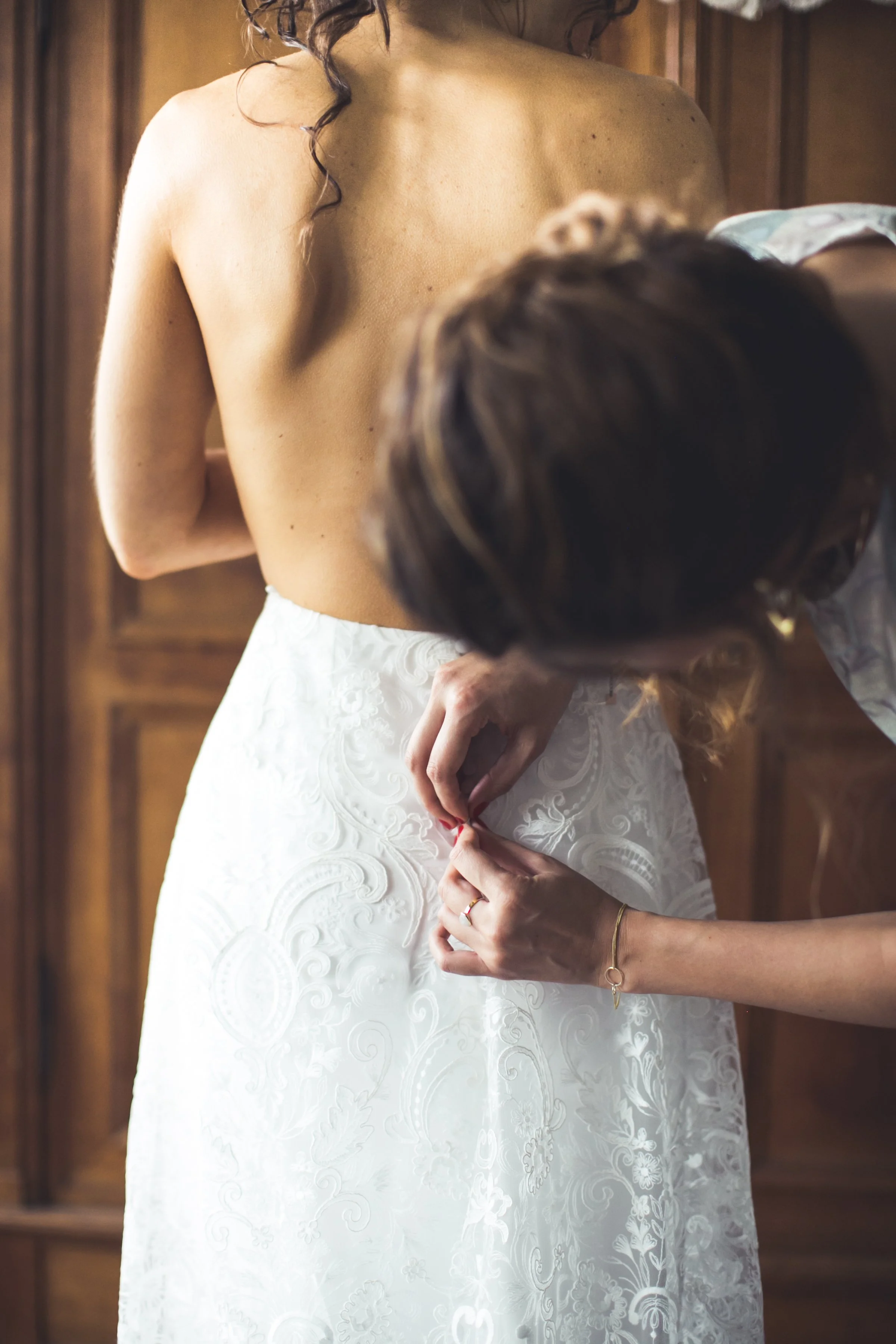 Een vrouw krijgt hulp terwijl ze haar witte bruidsjurk aandoet in een kamer met houten meubels op de achtergrond.