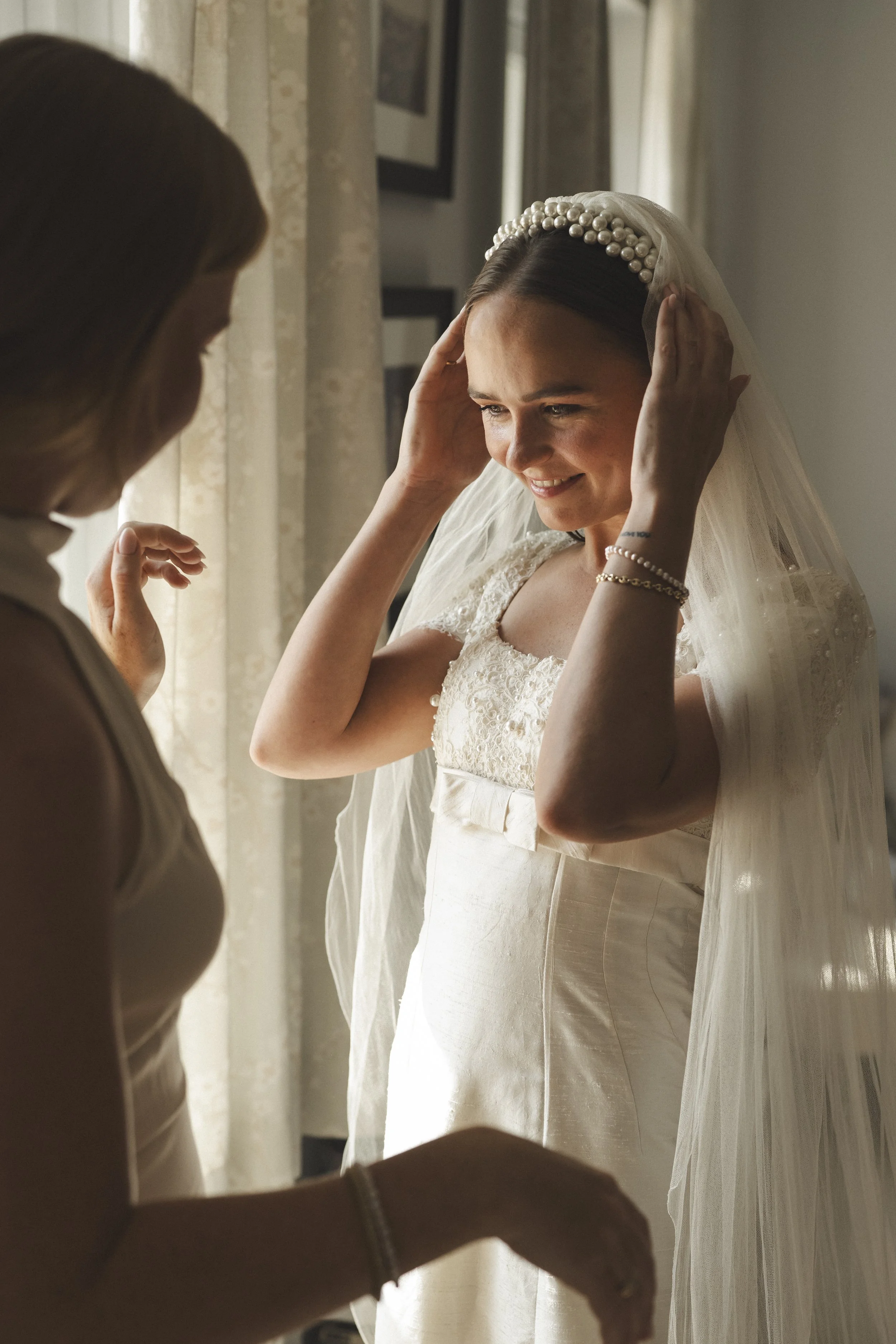 Een bruid die zich klaarmaakt op haar trouwdag terwijl een vriendin of familielid haar helpt. Ze kijkt in de spiegel en lacht.