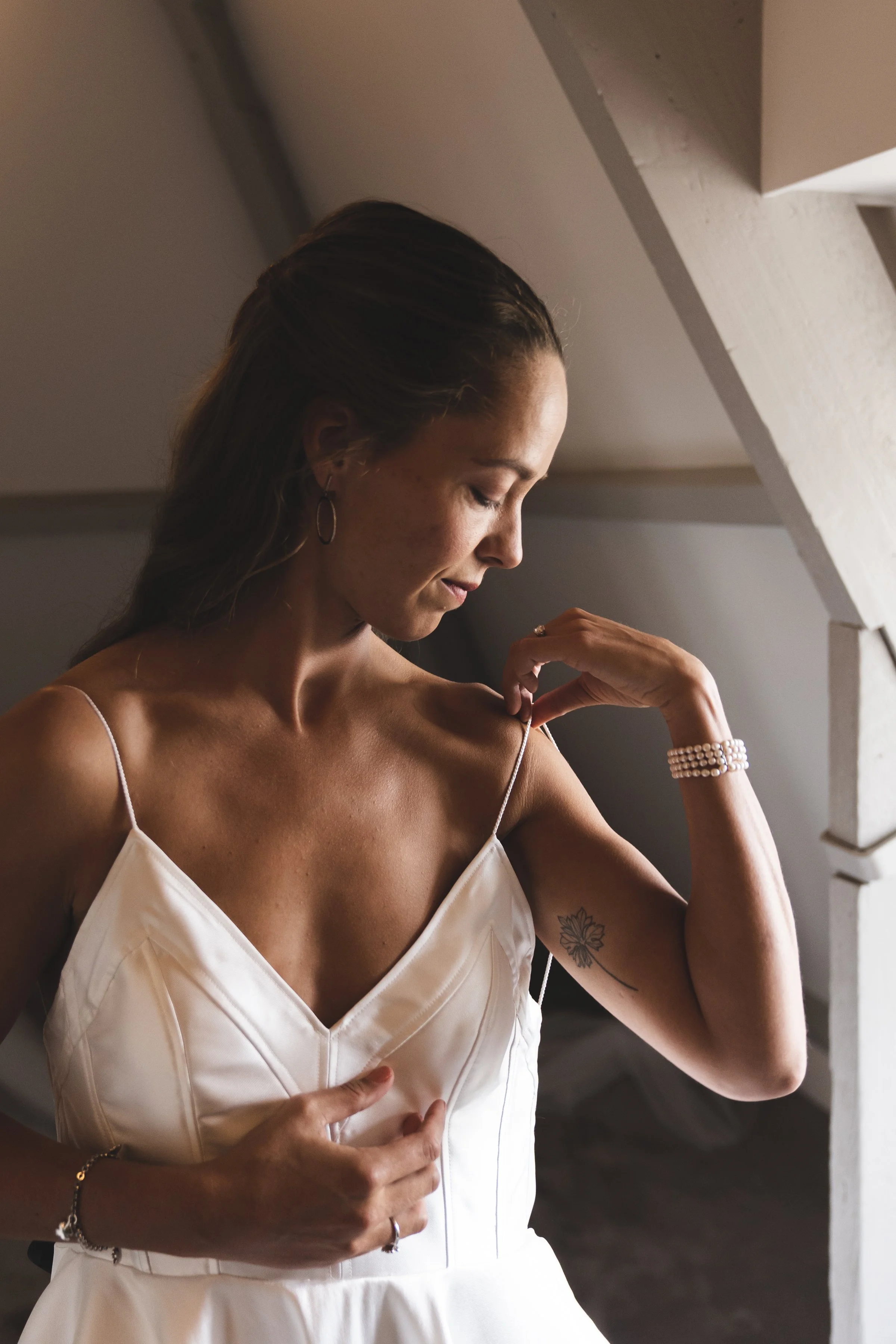 Een vrouw in een witte jurk die haar schouder aanraakt, met een tattoo op haar linkerarm, in een vredige binnenruimte.