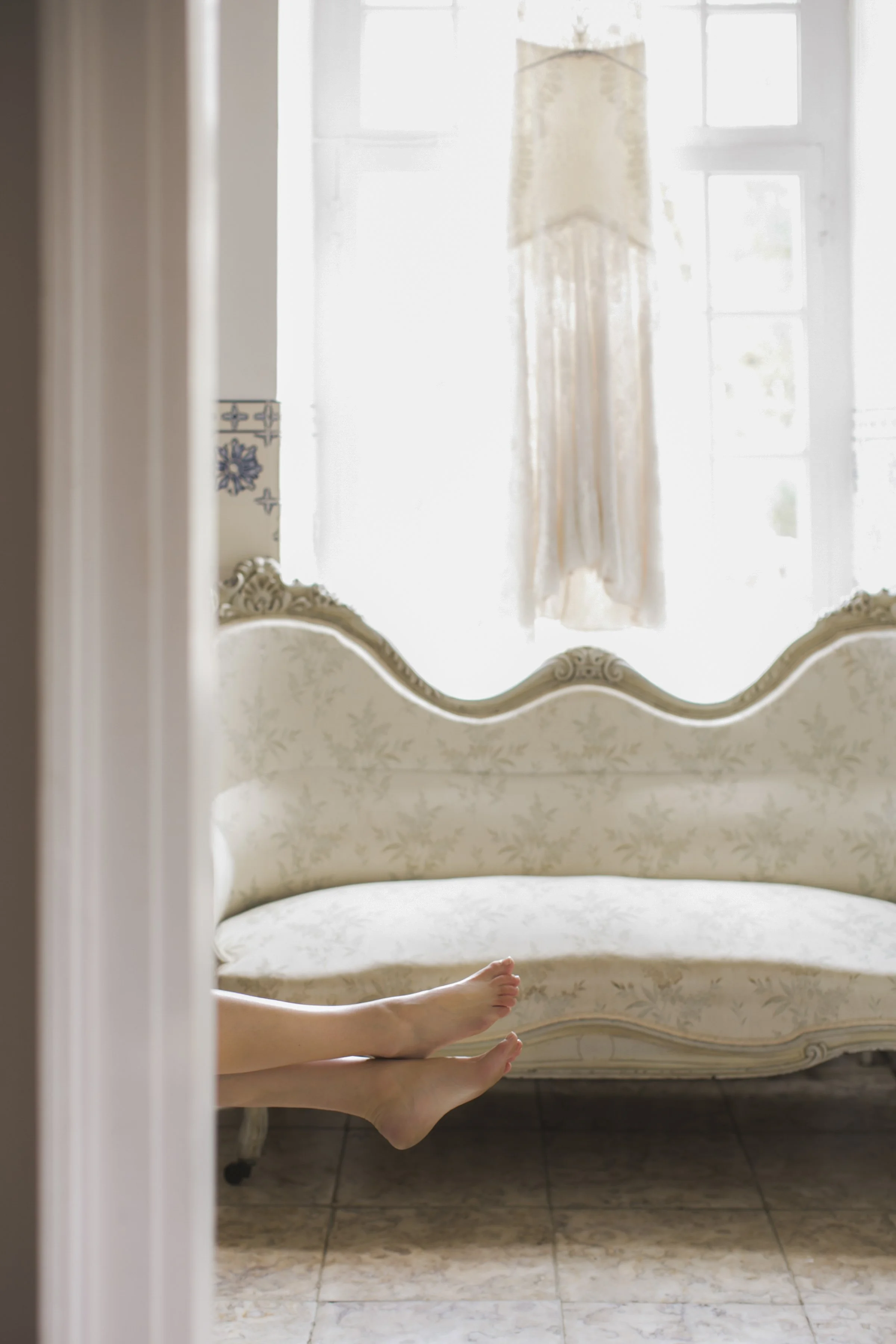 Naakte benen liggen op een vintage gestoffeerd bankje in een lichte kamer, met een raam op de achtergrond.