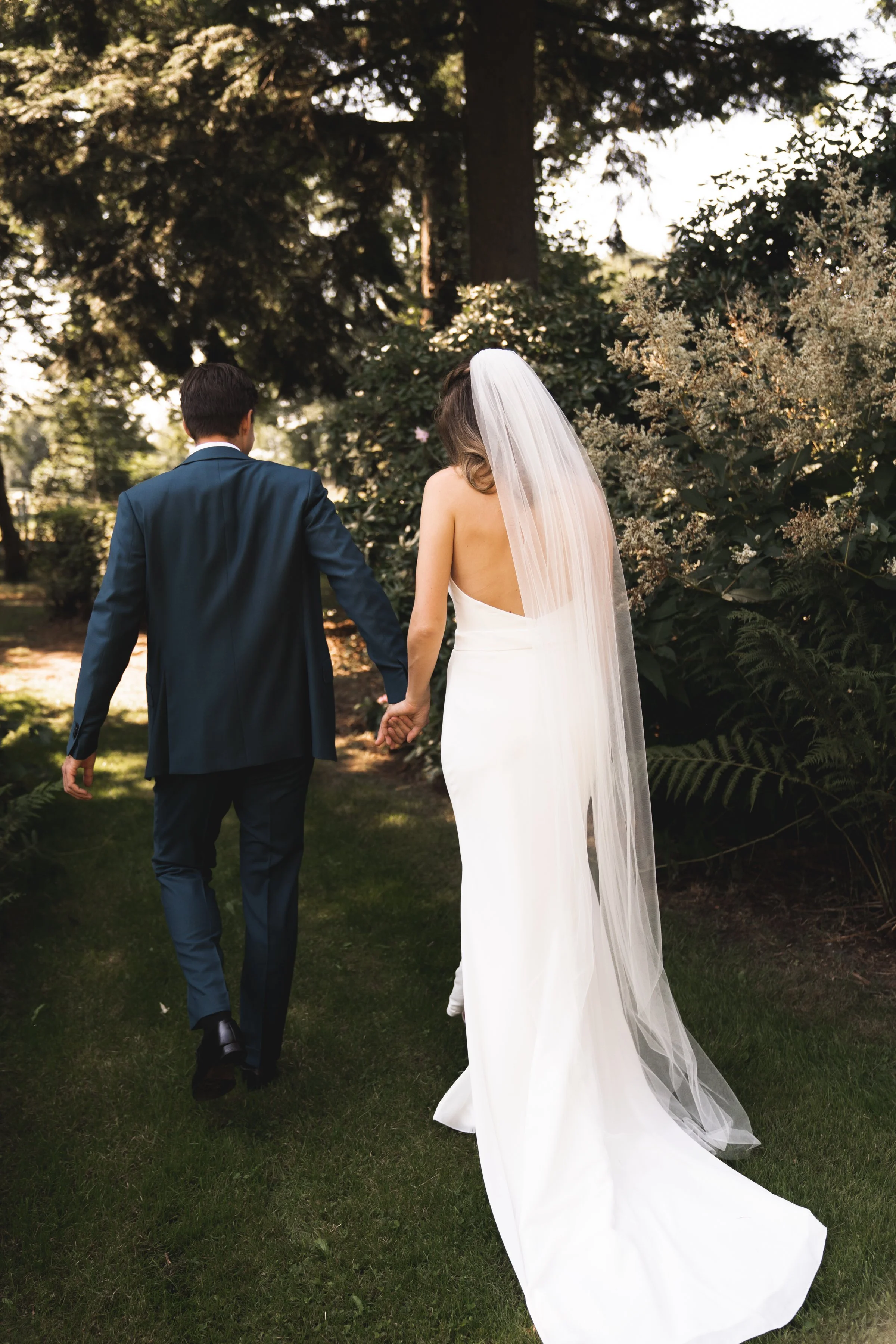 Een bruid en een bruidegom lopen hand in hand door een tuin, de bruid draagt een witte trouwjurk en een sluier, de bruidegom draagt een donkerblauwe pak.