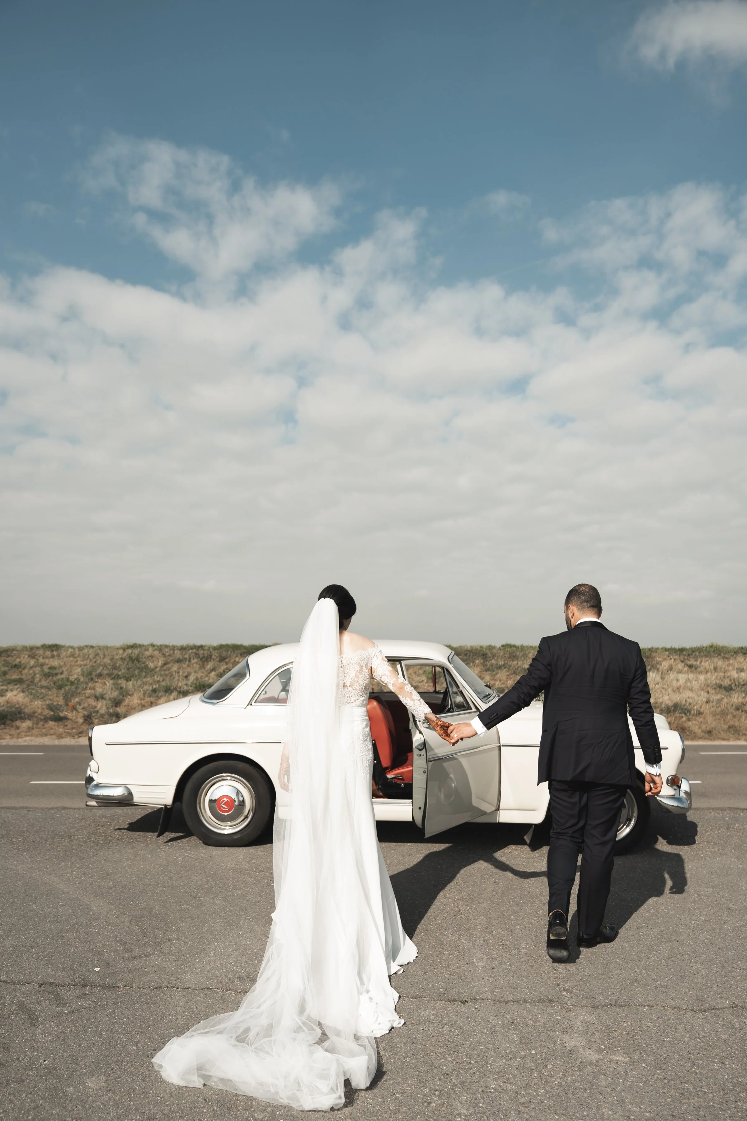 Een bruid en bruidegom lopen hand in hand weg van een witte klassieke auto op een open weg met een bewolkte blauwe lucht.