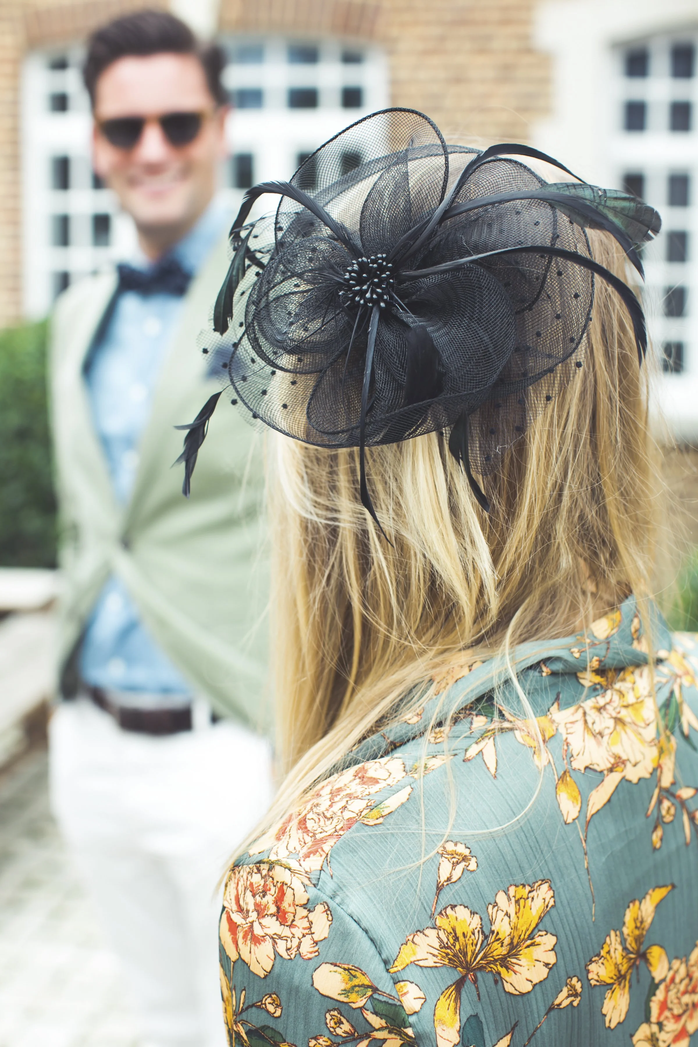 Close-up van een vrouw met lang blond haar die een grote, zwarte fascinator met veren draagt, terwijl een man in de achtergrond vlakbij staat, lachend, voor een gebouw met ramen.