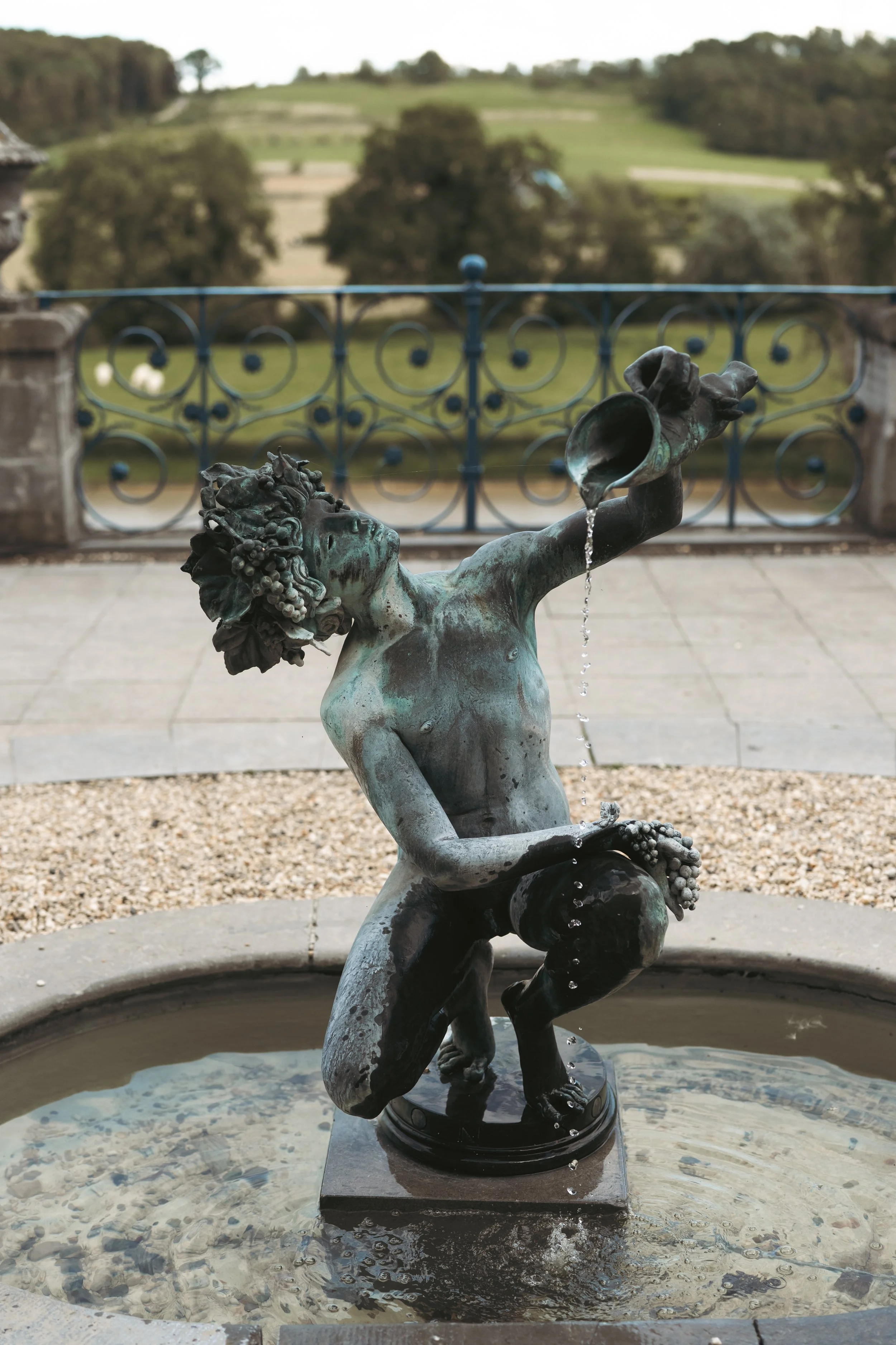 Bronzen beeld van een jongen die water uit een kruik drinkt, in een fontein, met een landelijke achtergrond.