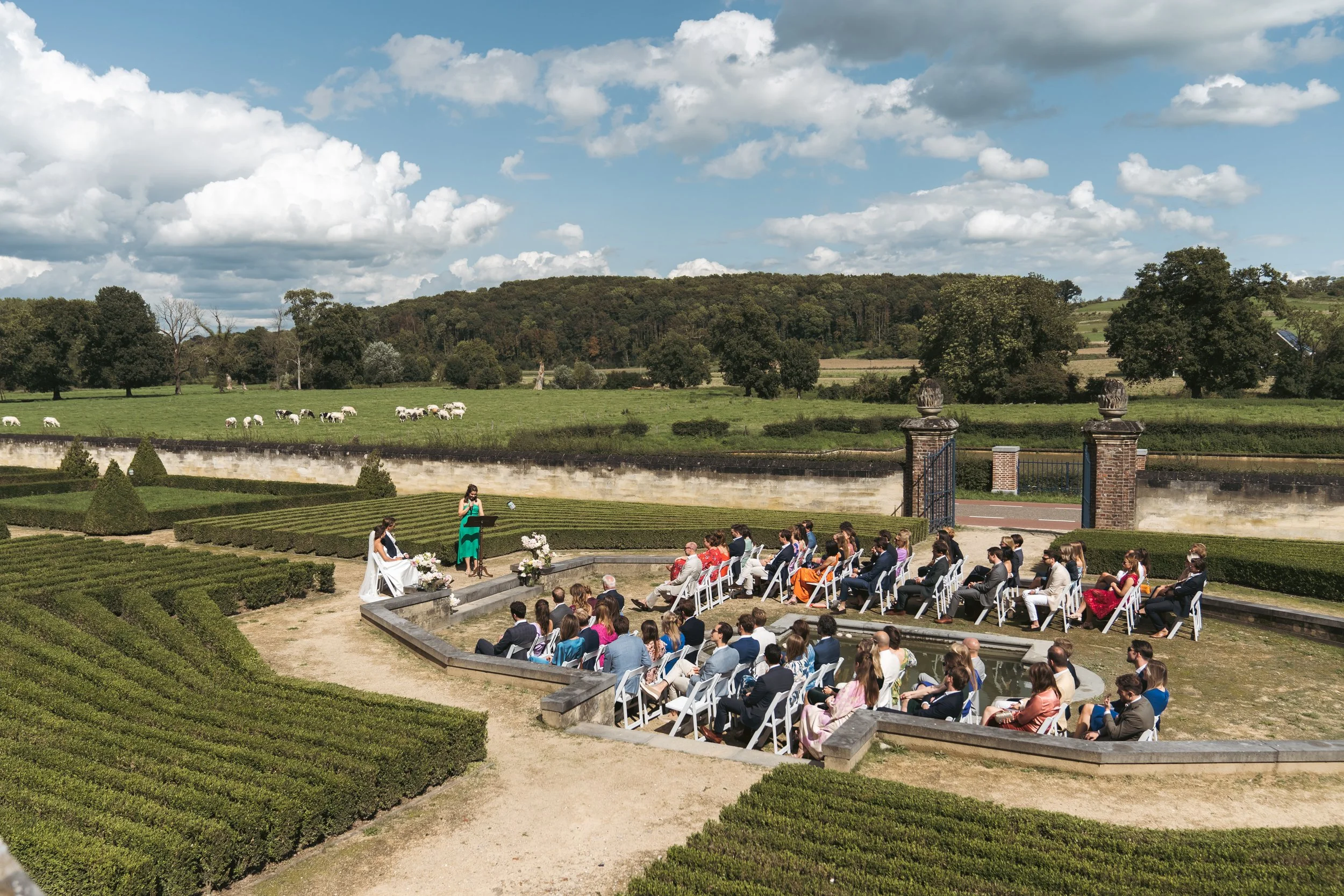 Buitense trouwceremonie in een tuin, met genodigden zittend op witte stoelen en een vrouw die ja zegt tegen een man, terwijl een vrouwelijke officiant spreekt. Op de achtergrond liggen groene velden en bomen onder een bewolkte hemel.