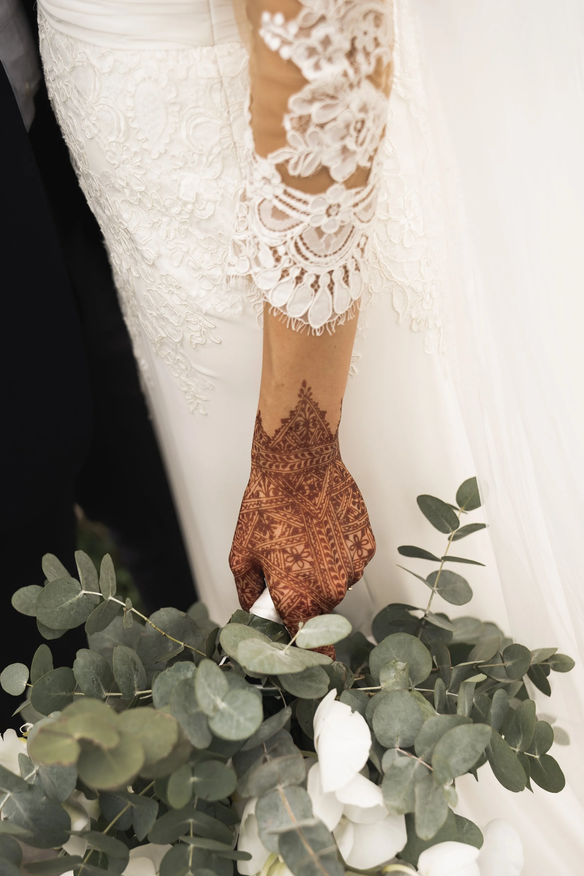 Close-up van een bruid in een witte jurk met kanten details, die een tak eucalyptus plukt of plaatst, met een getatoeëerde arm.