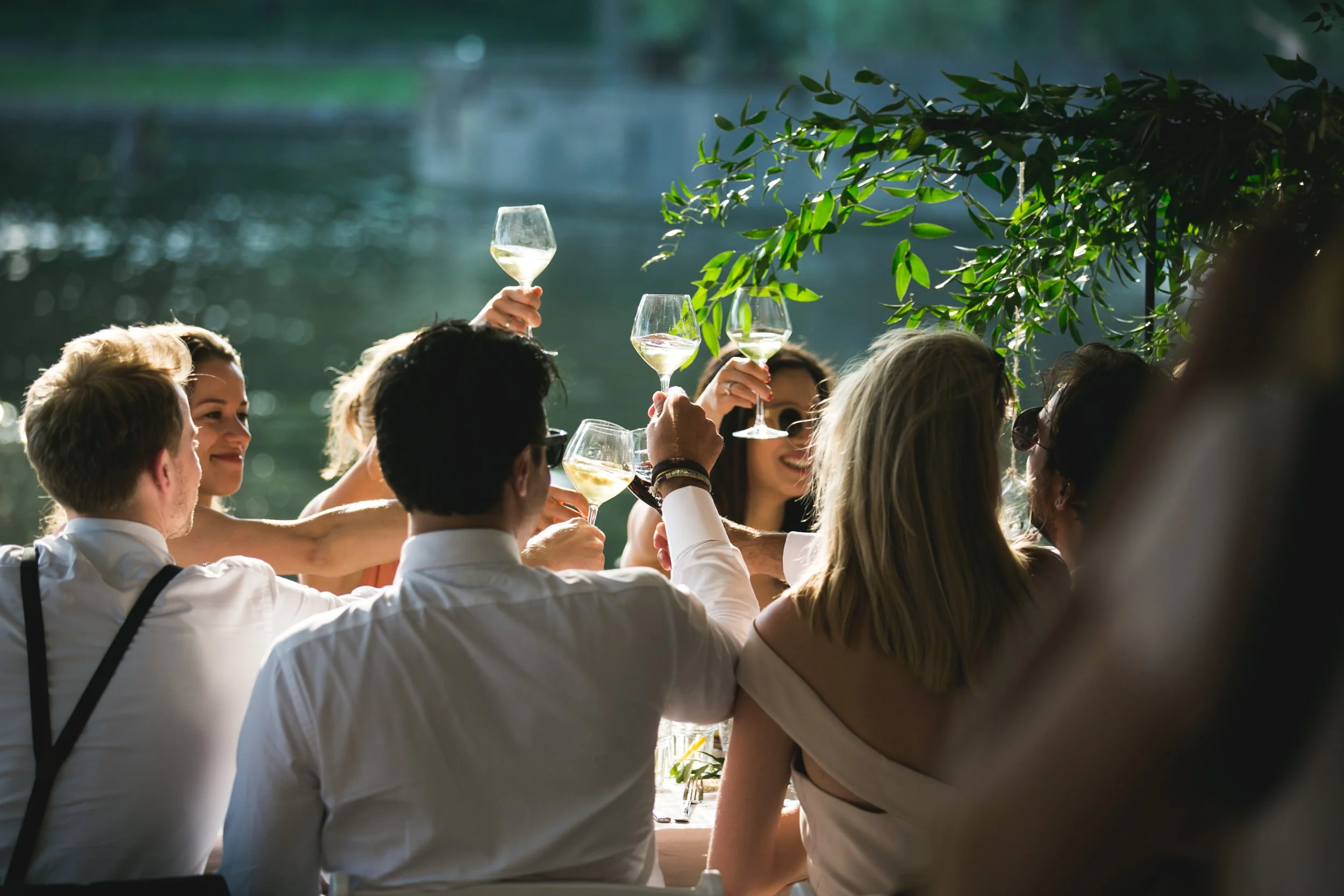 Mensen die samen proosten met witte wijn aan een tafel bij een buitenbijeenkomst, met een waterachtergrond en groene bladeren.