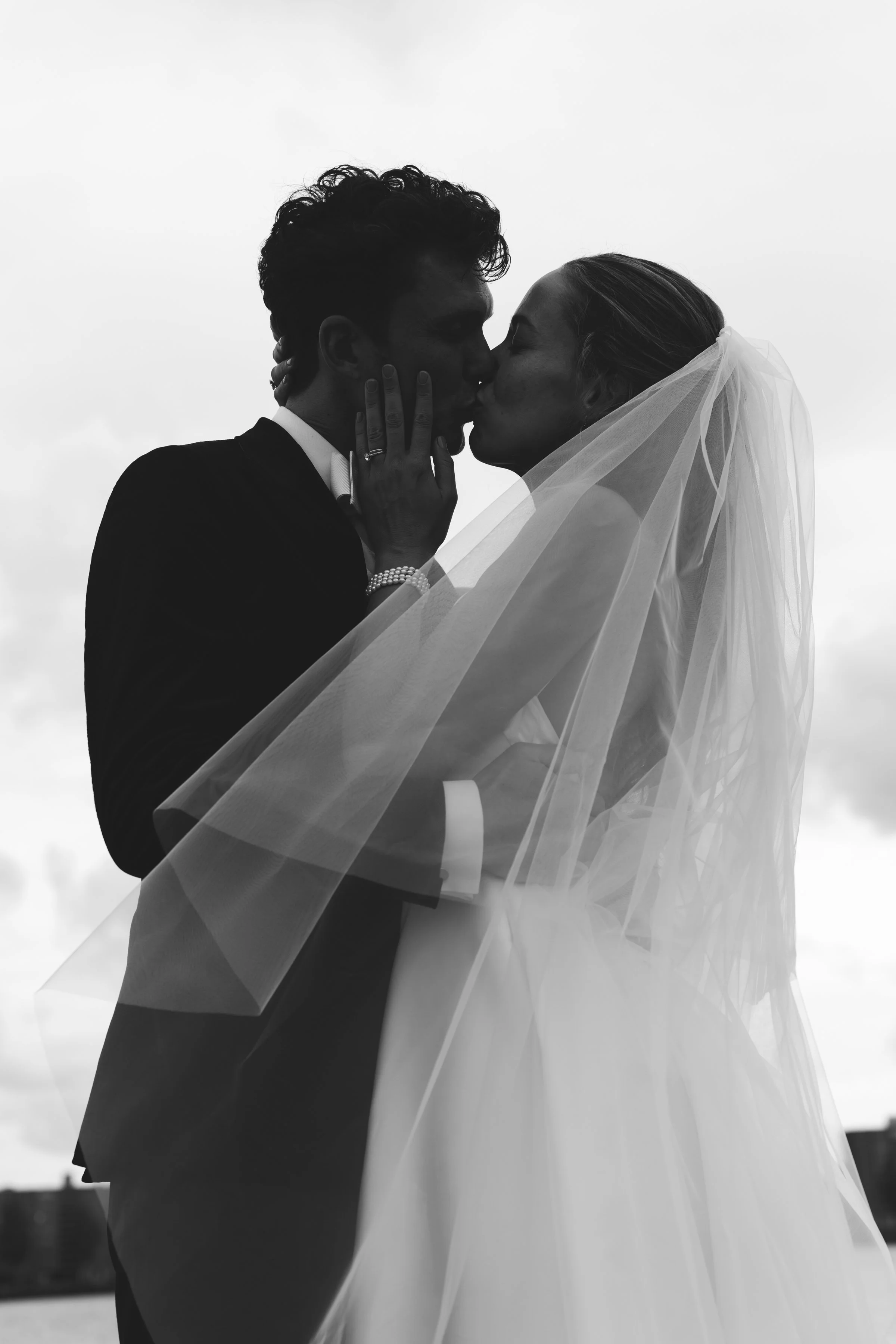 Twee mensen kussen, een van hen draagt een trouwjurk en sluier, de andere een pak, tegen een grijze hemel.