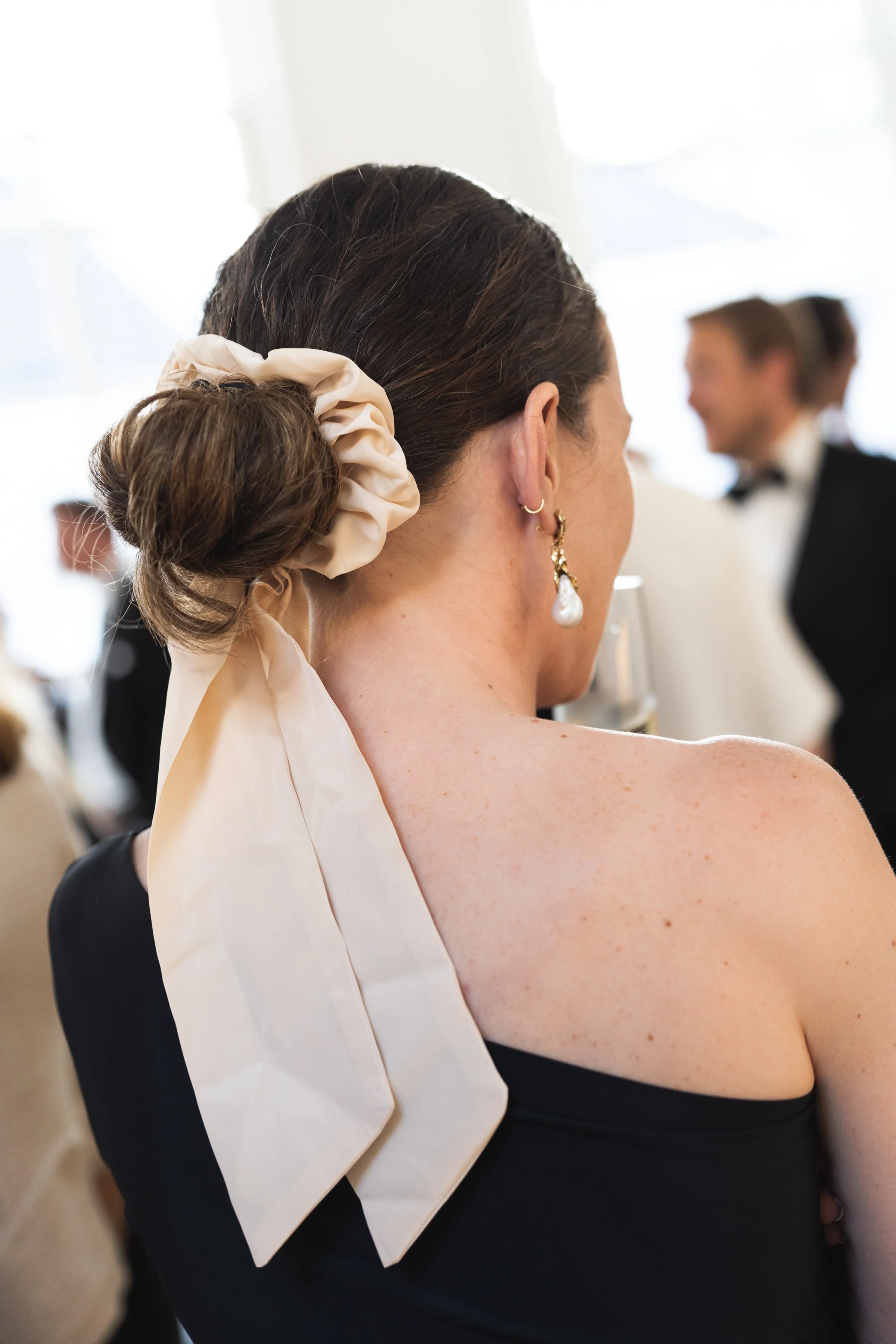Close-up van een vrouw met een chique haarstijl met een grote beige strik, die een pareloorbel draagt, tijdens een formeel evenement, met andere gasten op de achtergrond.