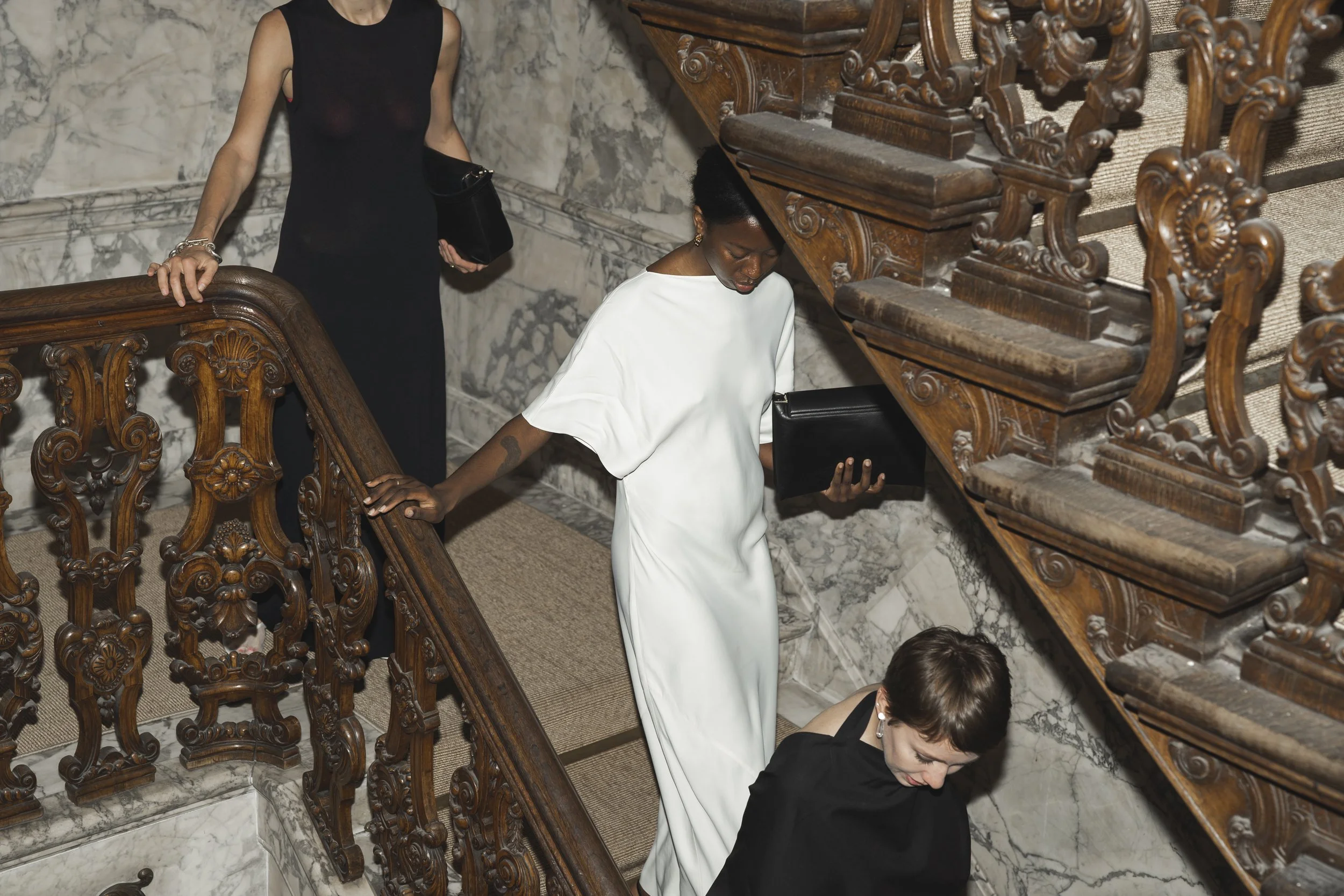 Drie vrouwen in formele kleding maken een trap in een gebouw met marmeren muren en een houten leuning.