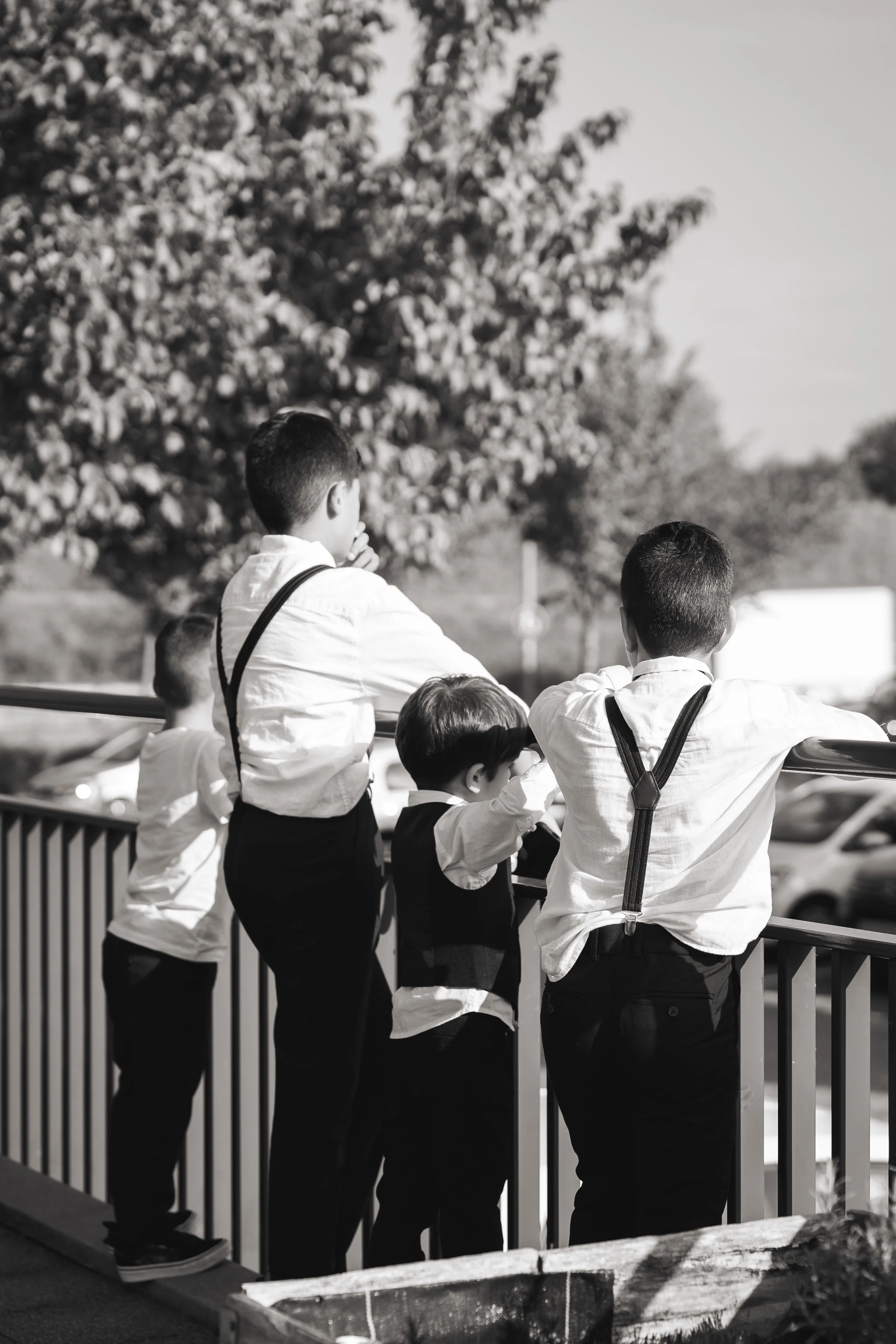 Vijf kinderen kijken vanuit een omheining, gekleed in formele kleding, op een zonnige dag met bomen op de achtergrond.