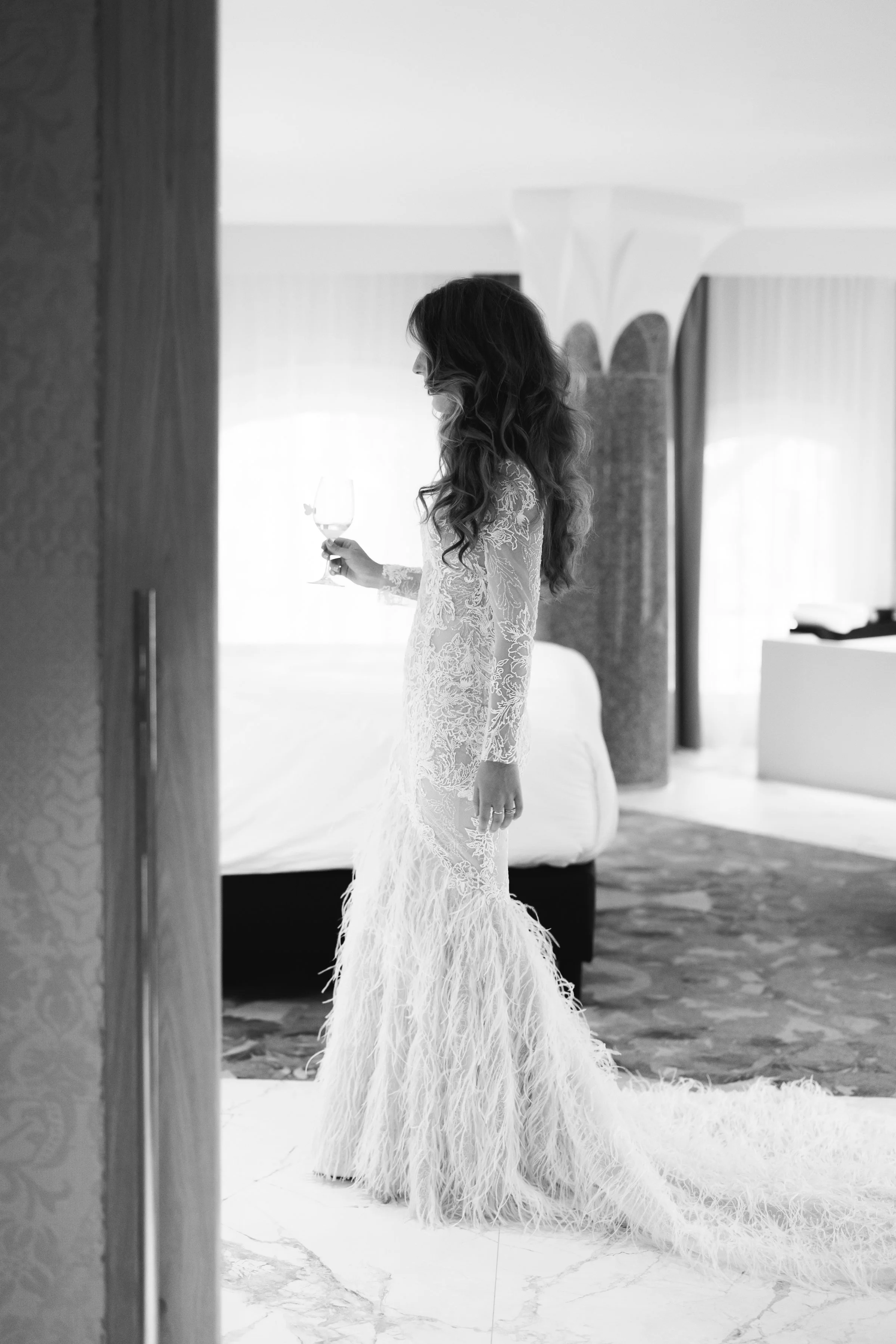Zwart-wit foto van een vrouw in een kanten trouwjurk met een lange, verenachtige rok, die naar haar wijst en in een wijnglas kijkt, in een hotelkamer met bed en gordijnen.