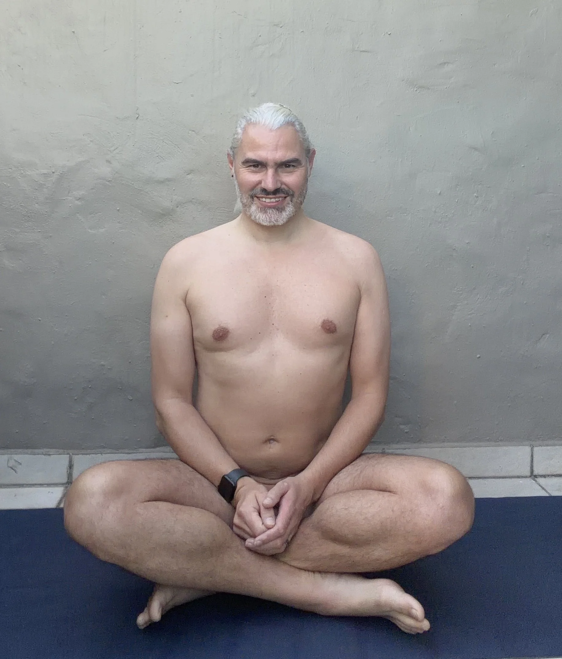 A smiling, middle-aged man with gray hair and a beard, sitting cross-legged on a yoga mat against a plain wall. He is shirtless and looking at the camera.