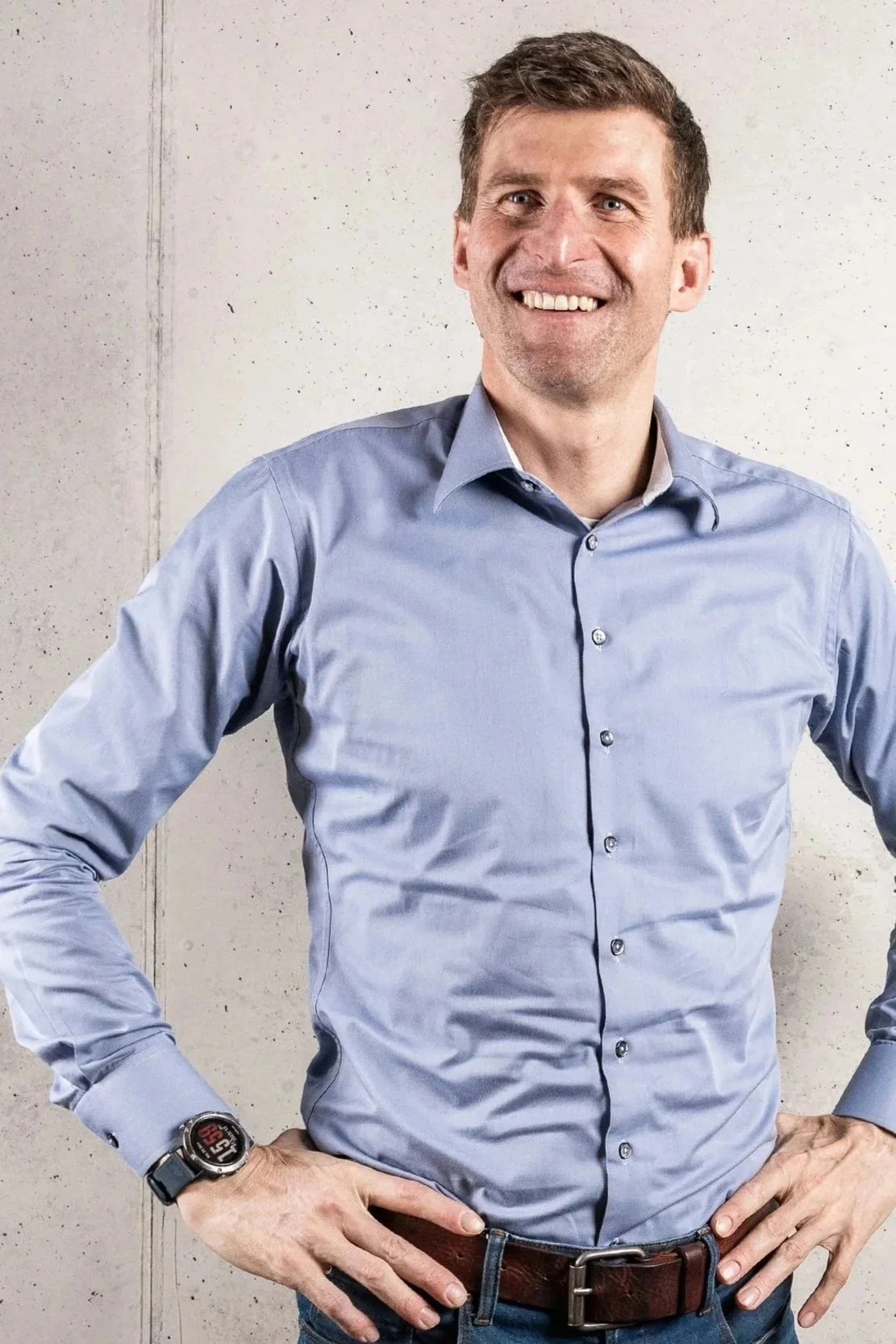 Ein Mann trägt ein hellblaues Hemd und eine Armbanduhr, steht vor einer Betonwand, lächelt und hat die Hände in die Hüften gestützt.
