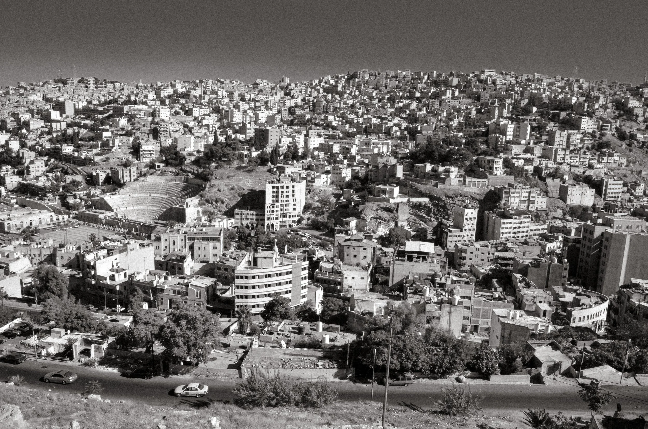 A black and white photo of a densely built hilly cityscape with numerous buildings, a stadium, and a street with cars in the foreground.