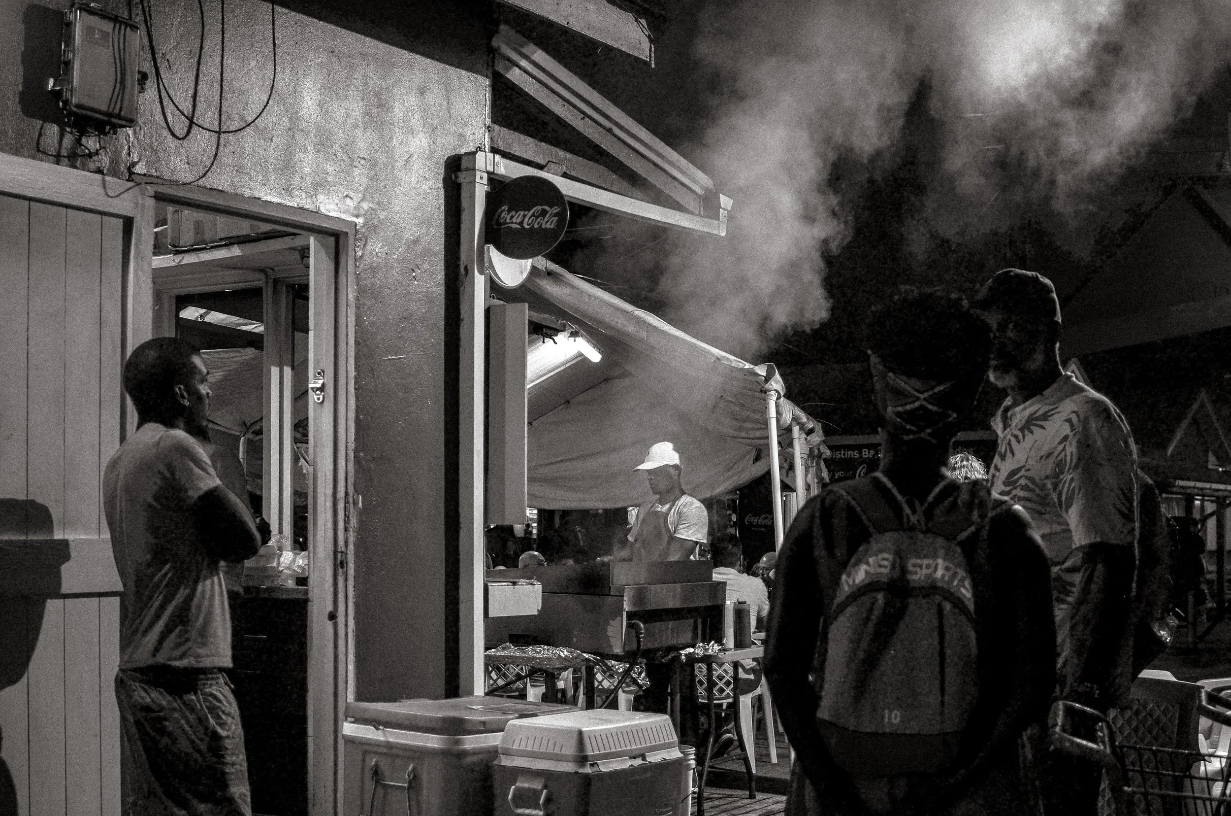 Grill smoke rises as people wait between stalls. Oistins, Barbados. December 2022.
