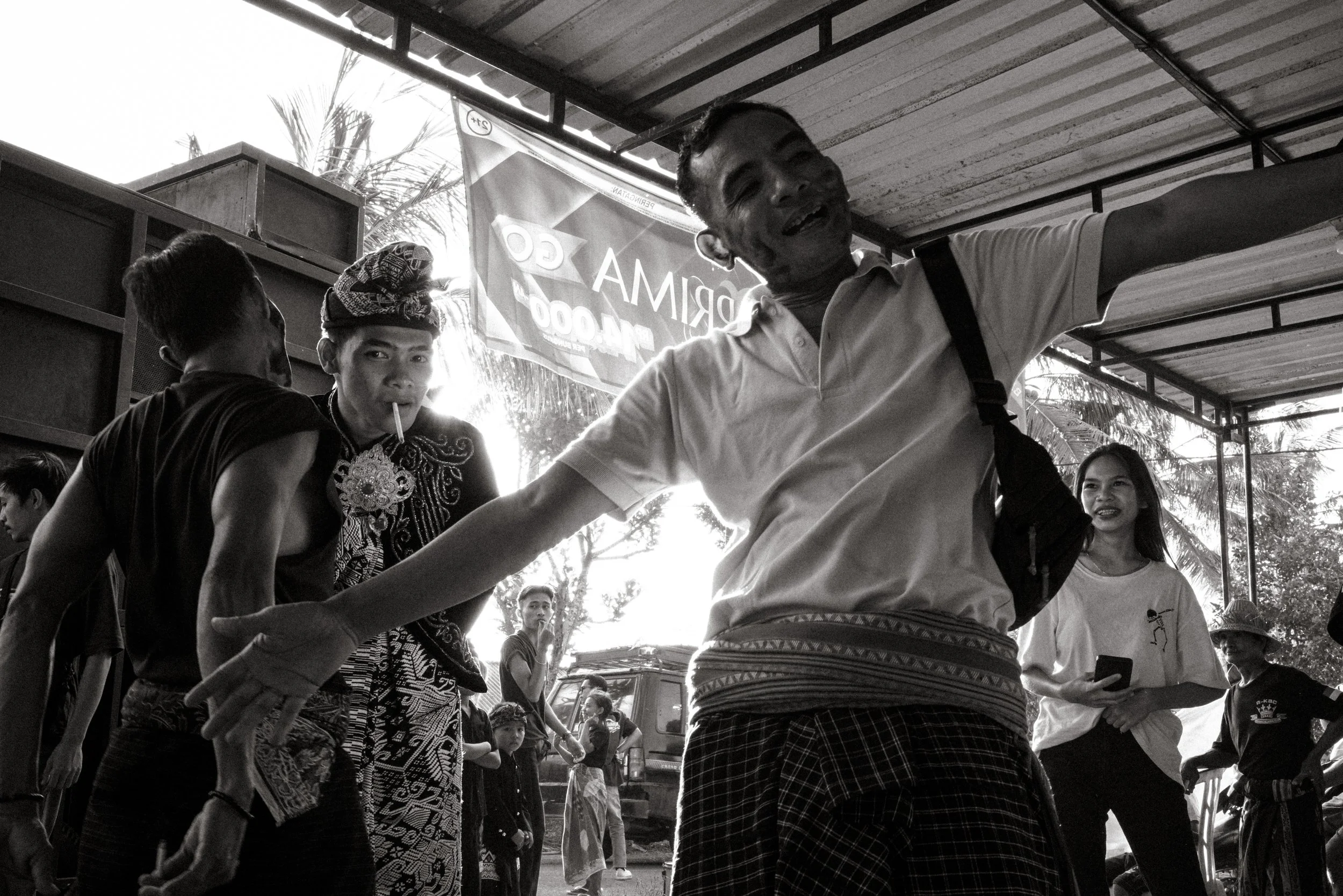 A group of people at an outdoor gathering, with one man prominently smiling and extending his arms outward, while others are engaged or observing, under a covered structure.