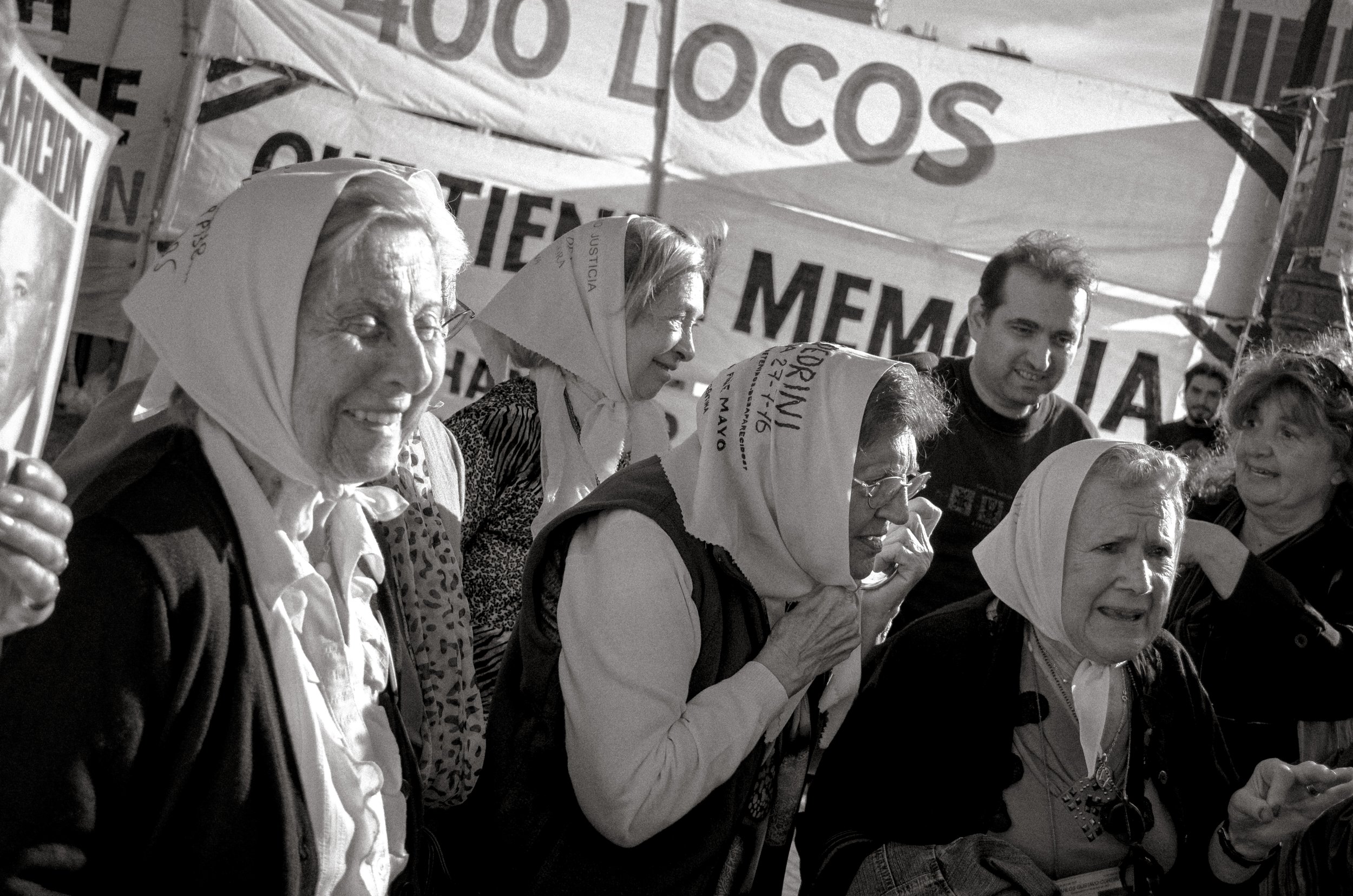 People protesting at a rally, some with headscarves, holding signs and banners, with a large banner in the background.