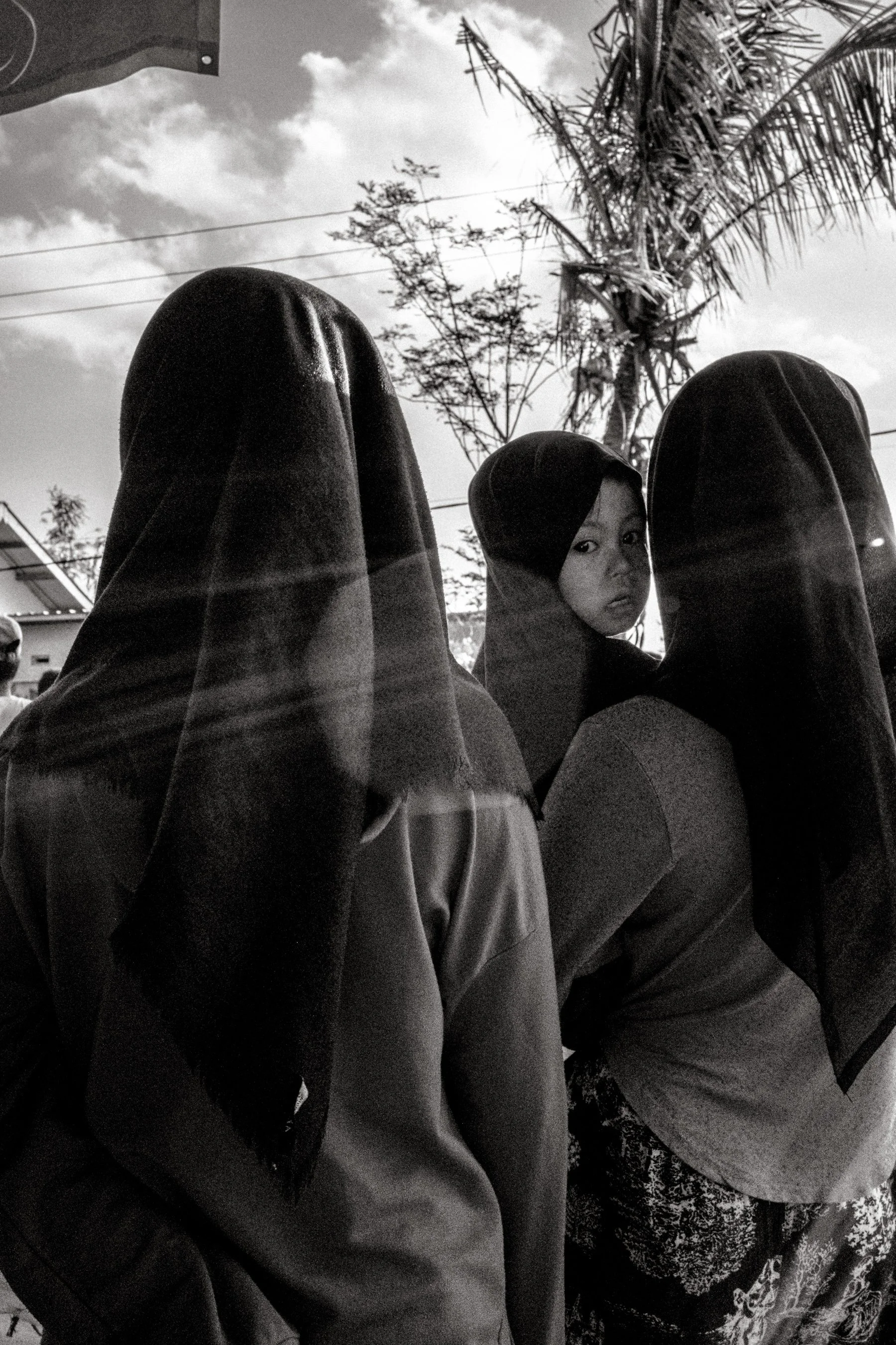 Women in headscarves stand together at the edge of the gathering as a girl looks back toward the camera