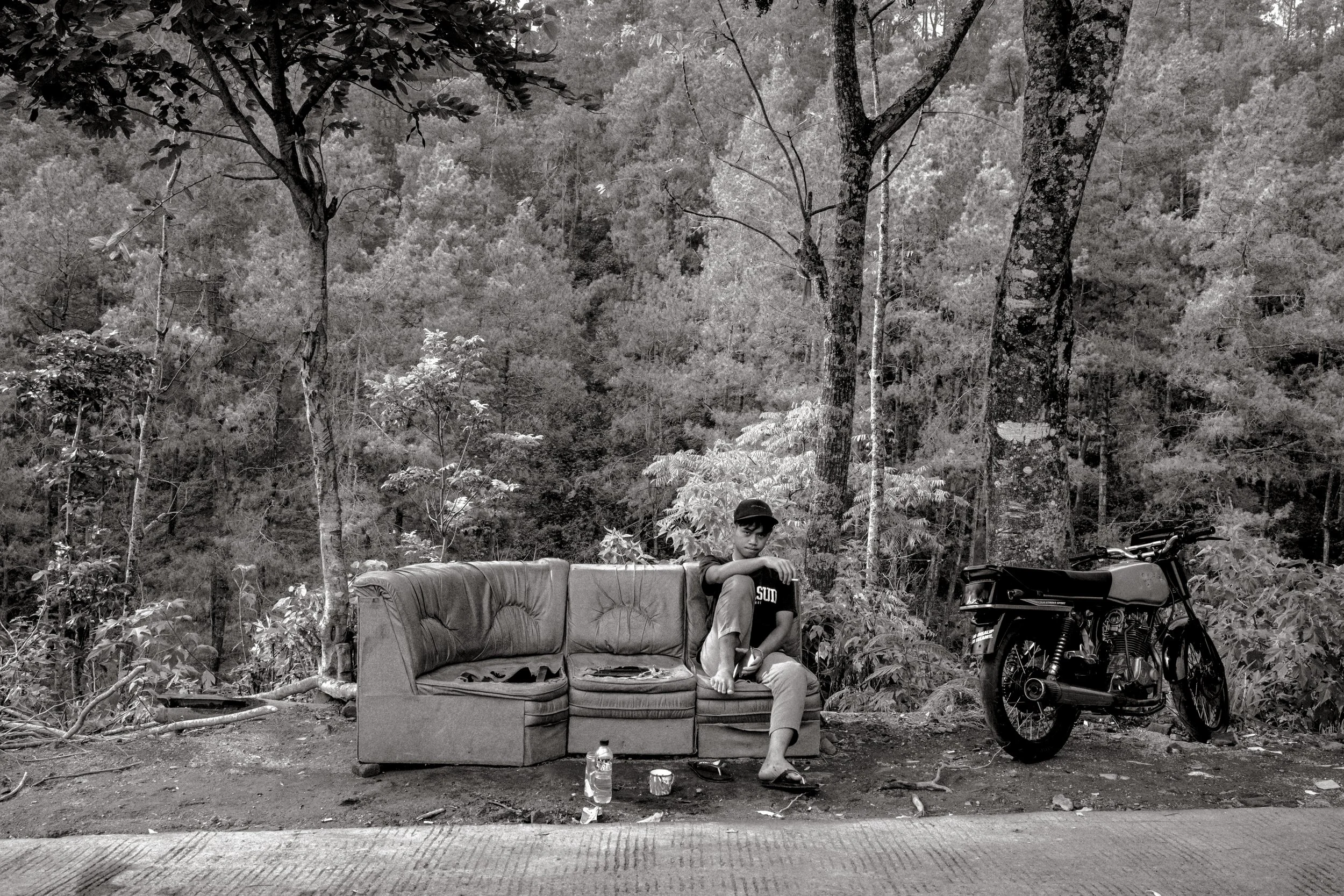 A man sitting on a worn sofa by the side of a forested road with a motorcycle parked next to him. The scene is in black and white.