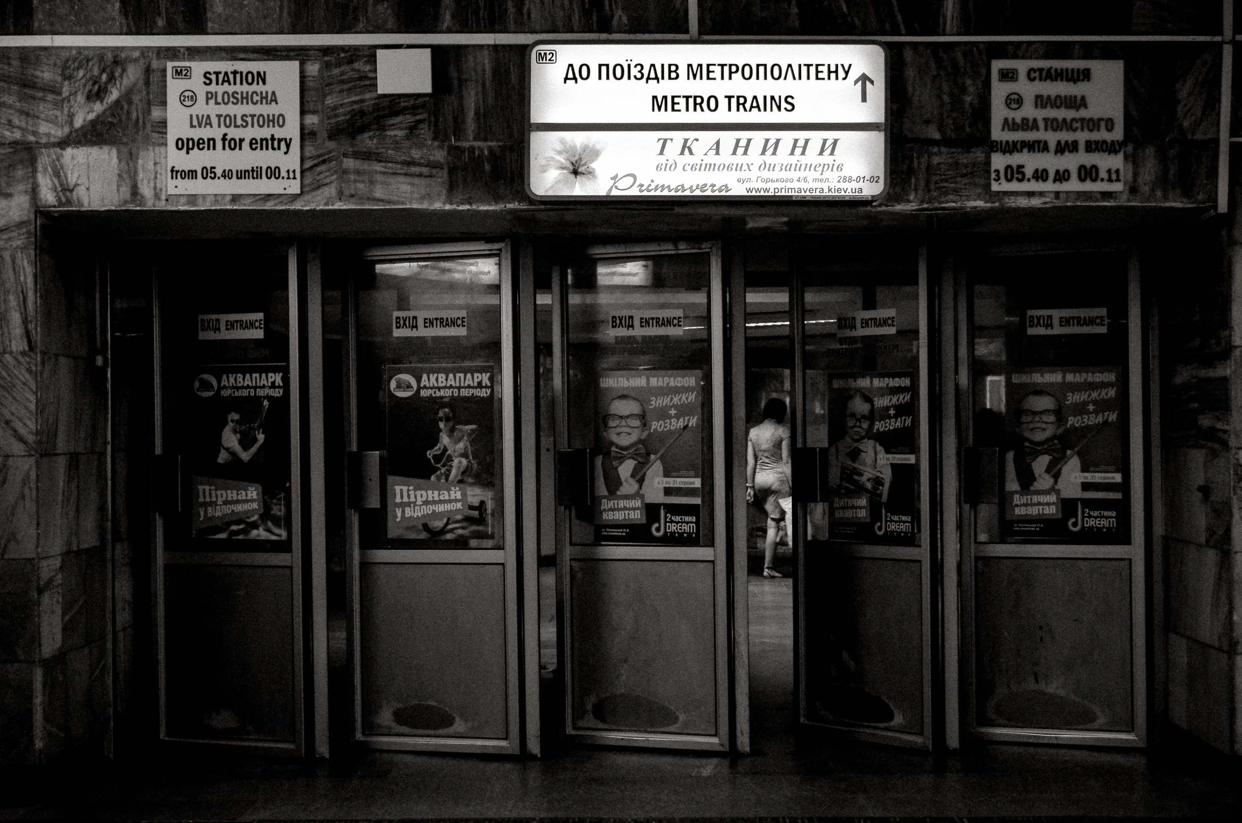 Entrance to a metro station with signage in Ukrainian and English, featuring advertisements and a person walking inside.